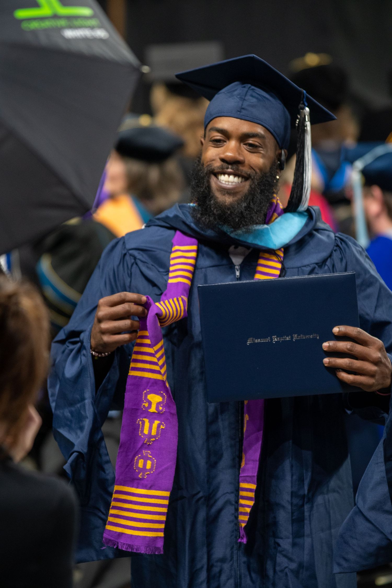 Graduate in blue regalia displays diploma while wearing colorful stoles at graduation ceremony.