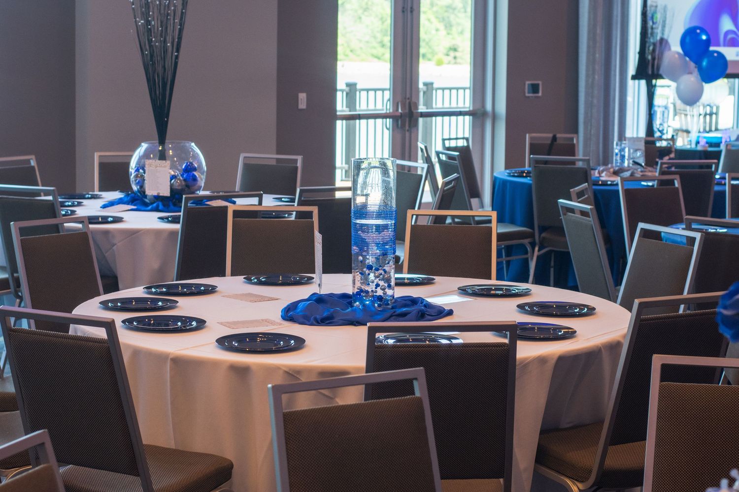 A formal banquet hall setup with round tables covered in white linens and blue centerpiece decorations.