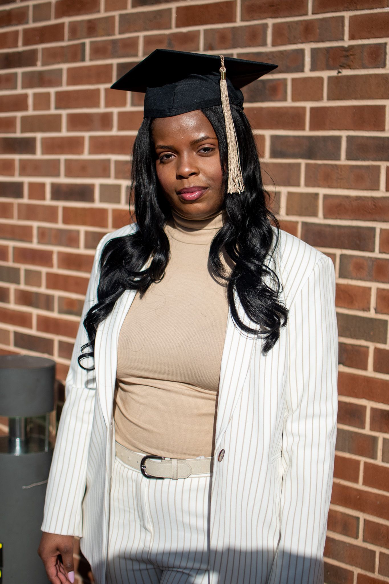 Graduate in academic regalia wearing a white pinstripe blazer and beige turtleneck poses against brick wall.