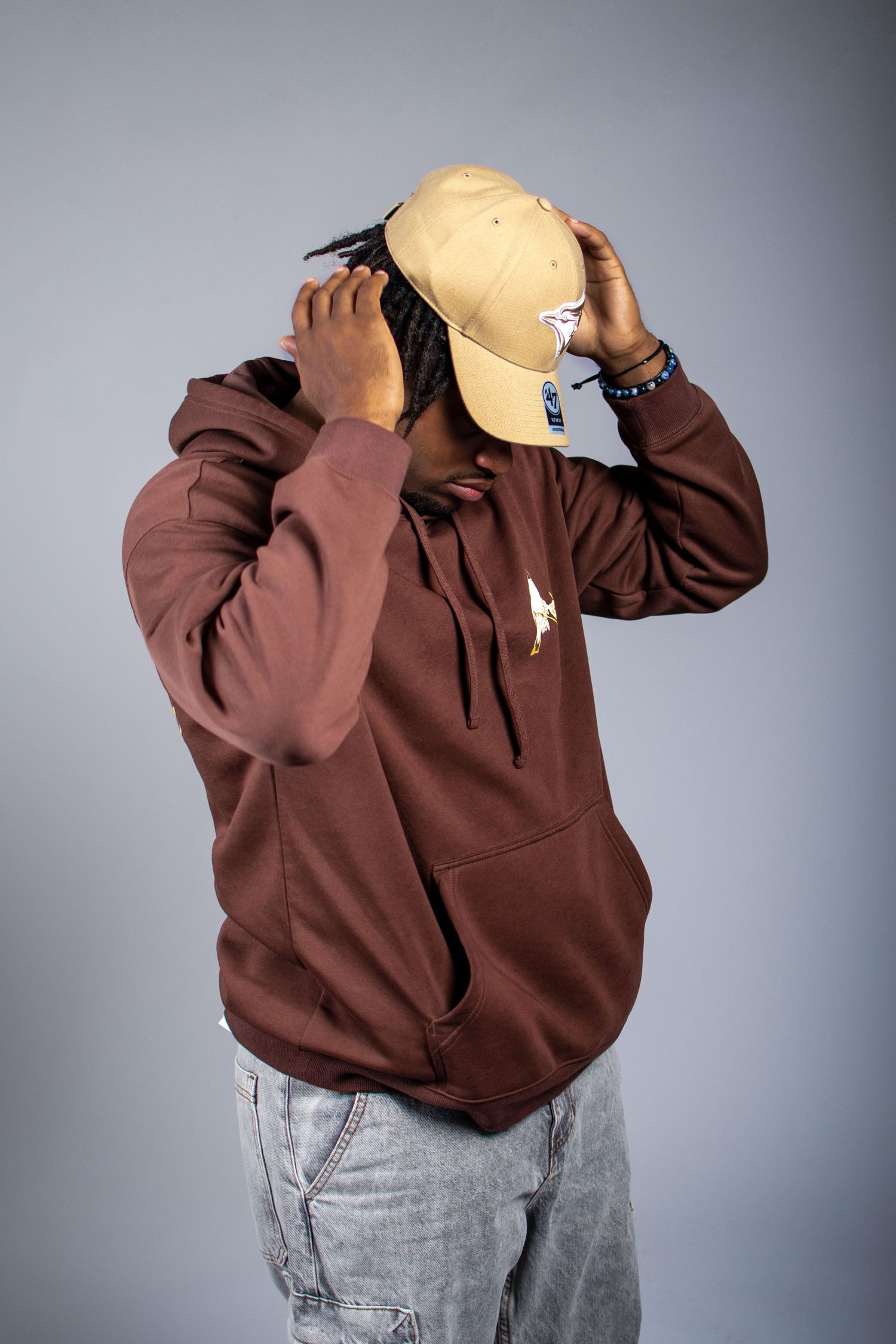 Person wearing a brown hoodie and beige baseball cap with light wash jeans against a gray background.
