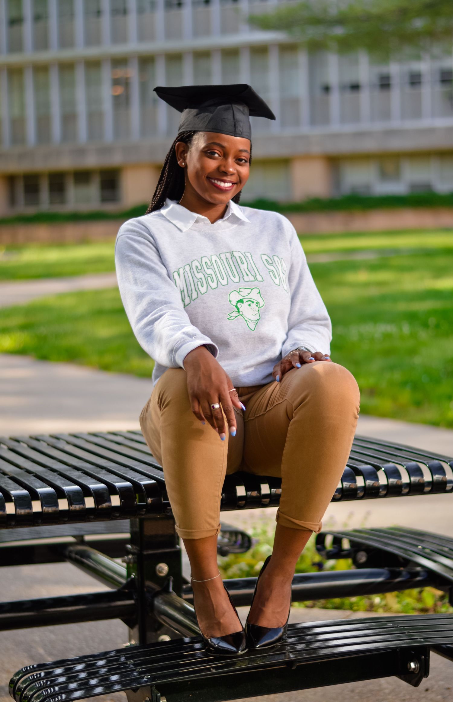 Graduate in cap and gown sitting on outdoor bench in casual pose.