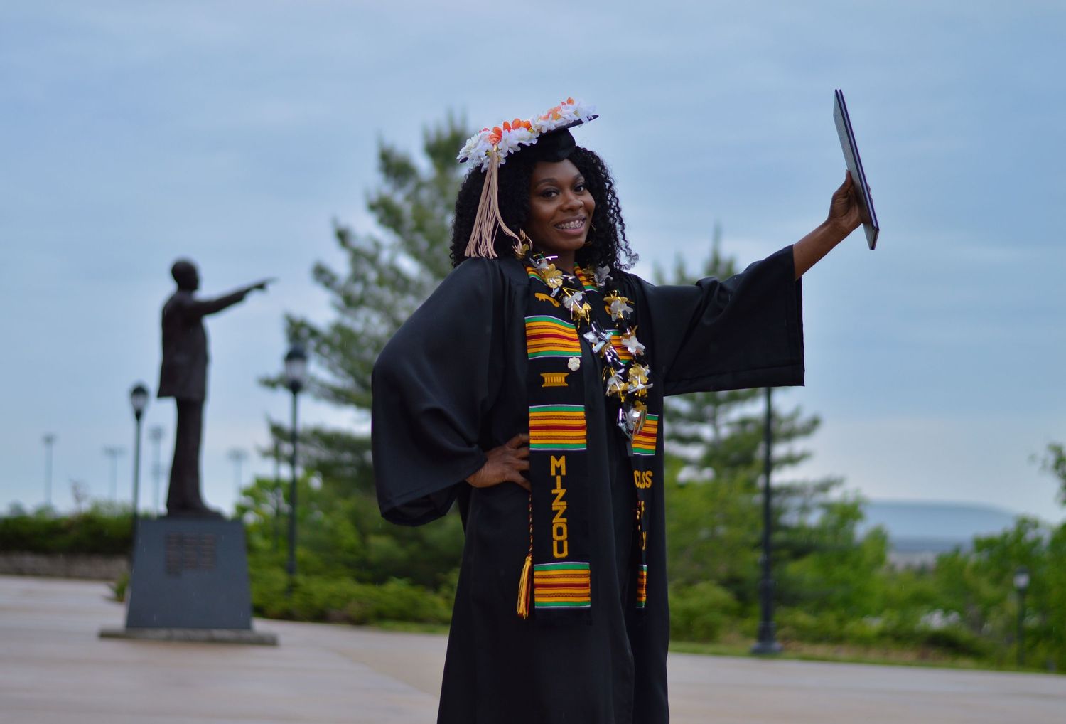Graduate in cap and gown with kente stole celebrates at outdoor campus location with statue silhouette in background.