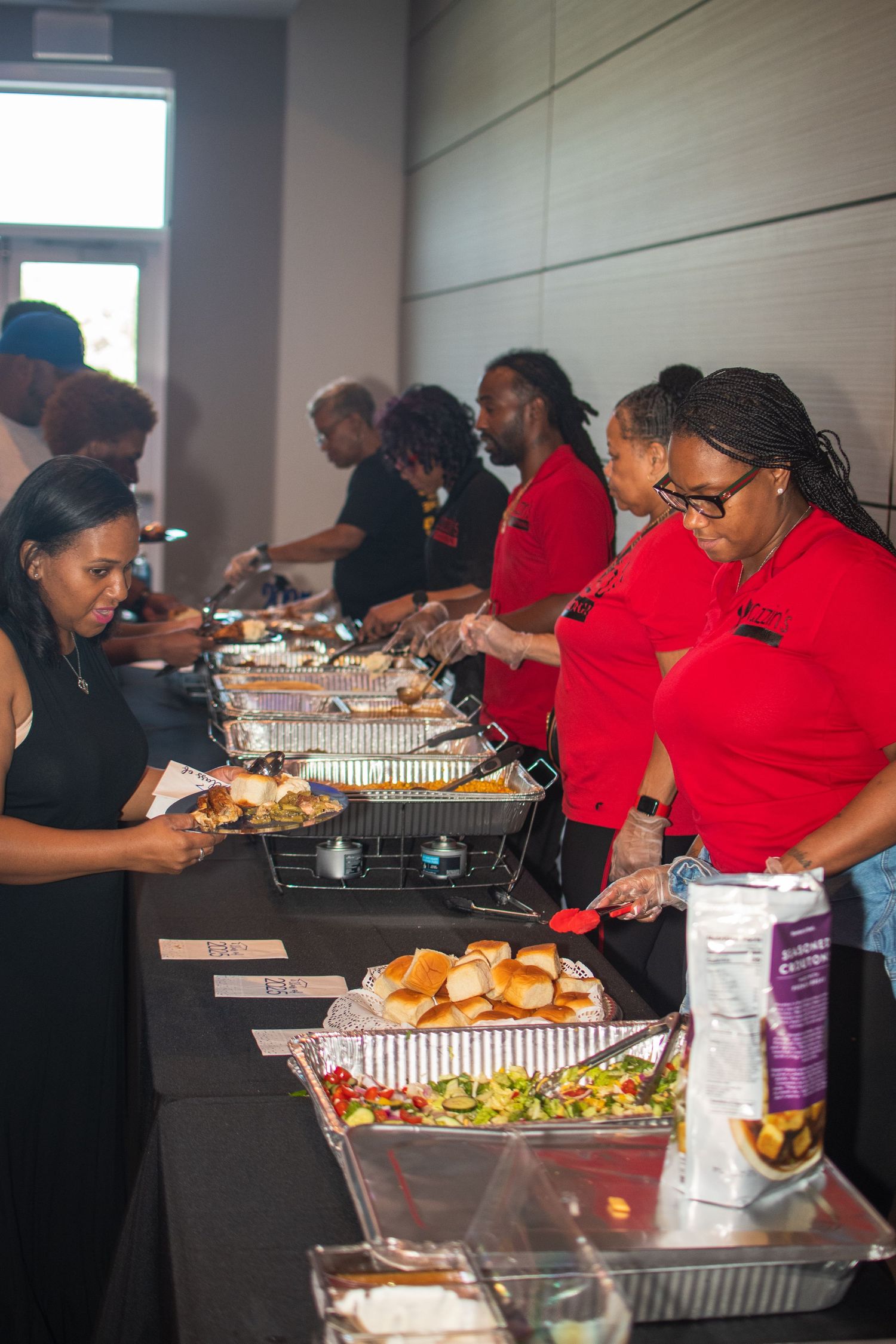 People in red shirts gather around a buffet table serving food at what appears to be a community or group meal event.