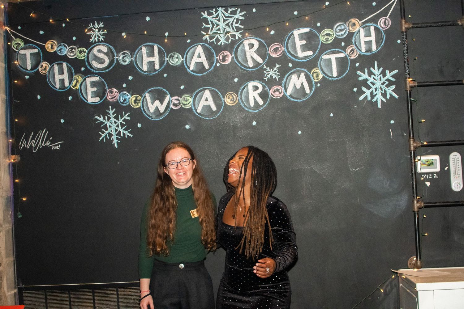 Students pose in front of a chalkboard decorated with snowflakes and holiday text during a winter celebration.