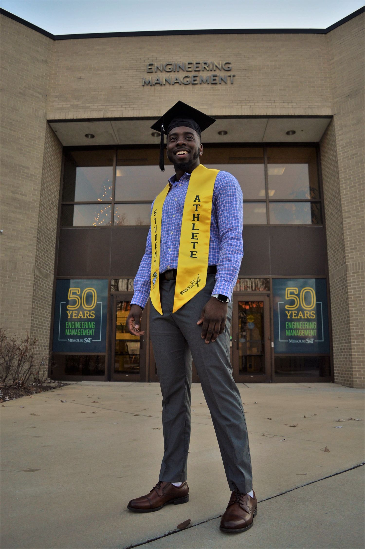 Graduate in business attire and honor cords poses outside engineering building.
