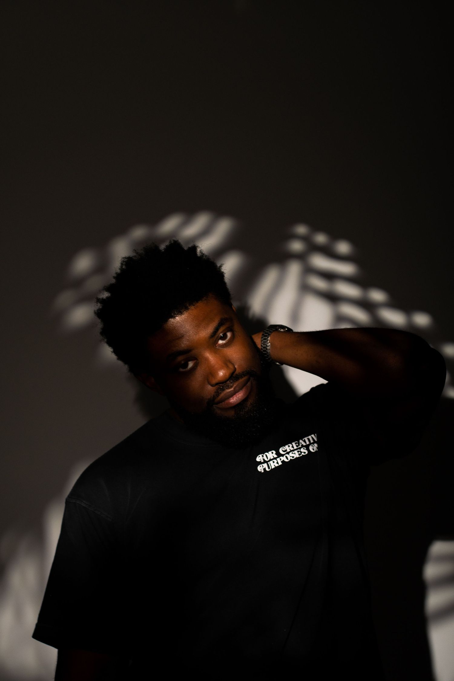 Young Black man in black graphic tee poses confidently against dark wall with dramatic shadow patterns.
