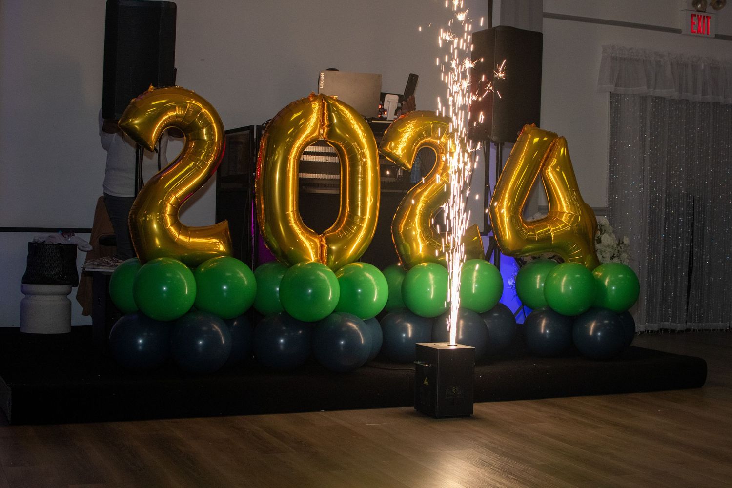 Gold metallic 2024 balloons with green and silver decorations and a sparkling fountain display in a dark room.
