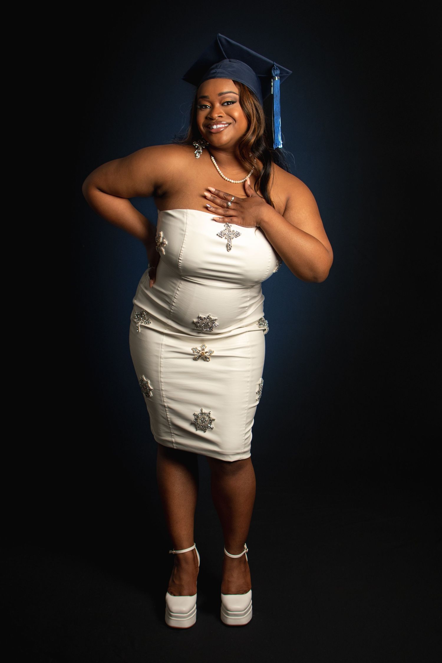 Creative low angle graduation portrait in white dress and navy cap against dark background.