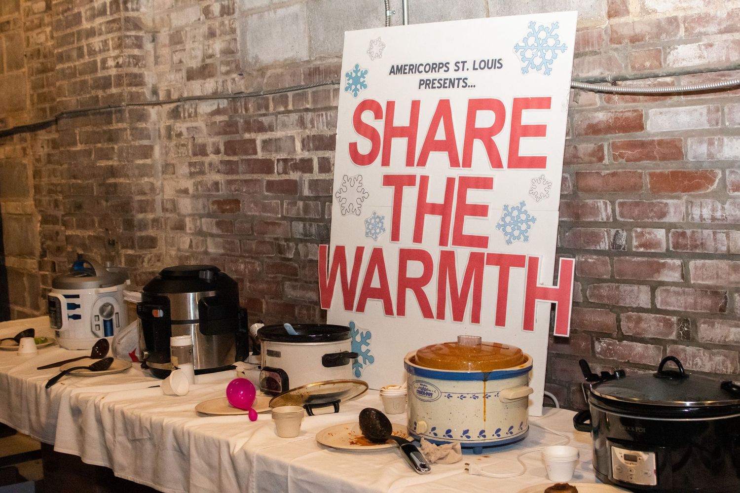 A 'Share the Warmth' sign hangs above a table with slow cookers and food service equipment against a brick wall.