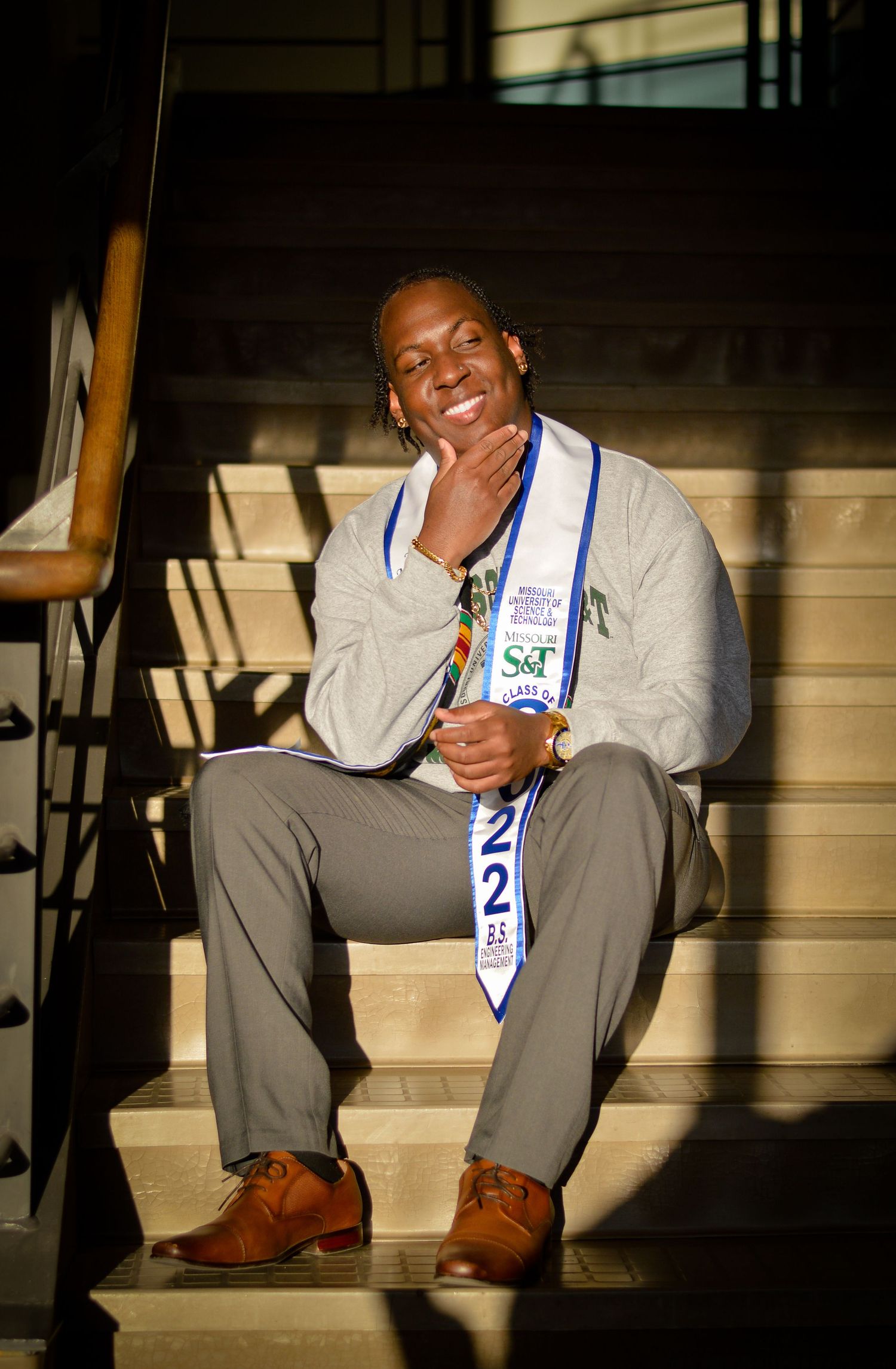 A person wearing a sash sits on stairs in business casual attire, smiling warmly in natural lighting.