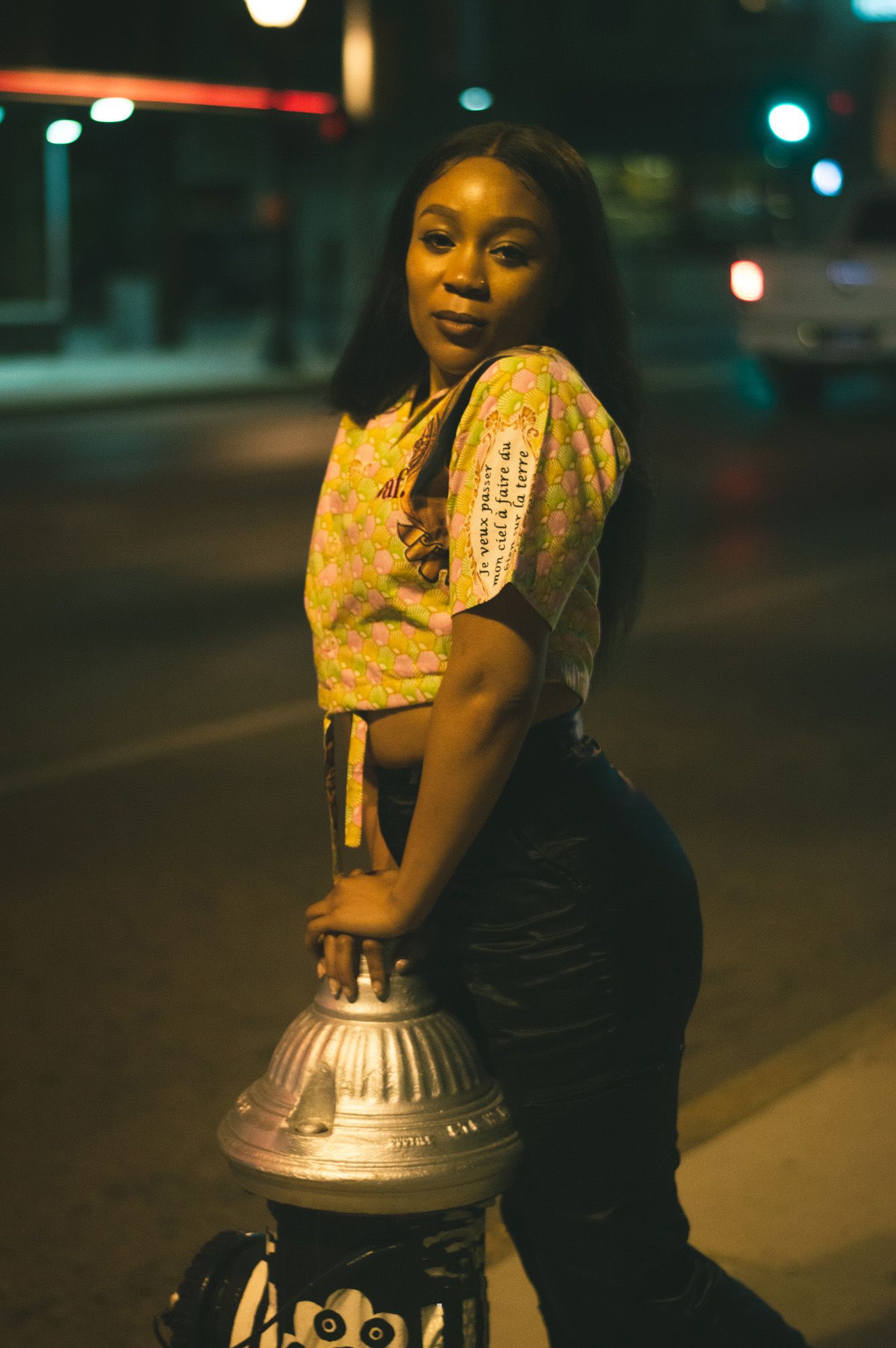 Person in yellow patterned top and dark pants poses against silver fire hydrant on city street at night.