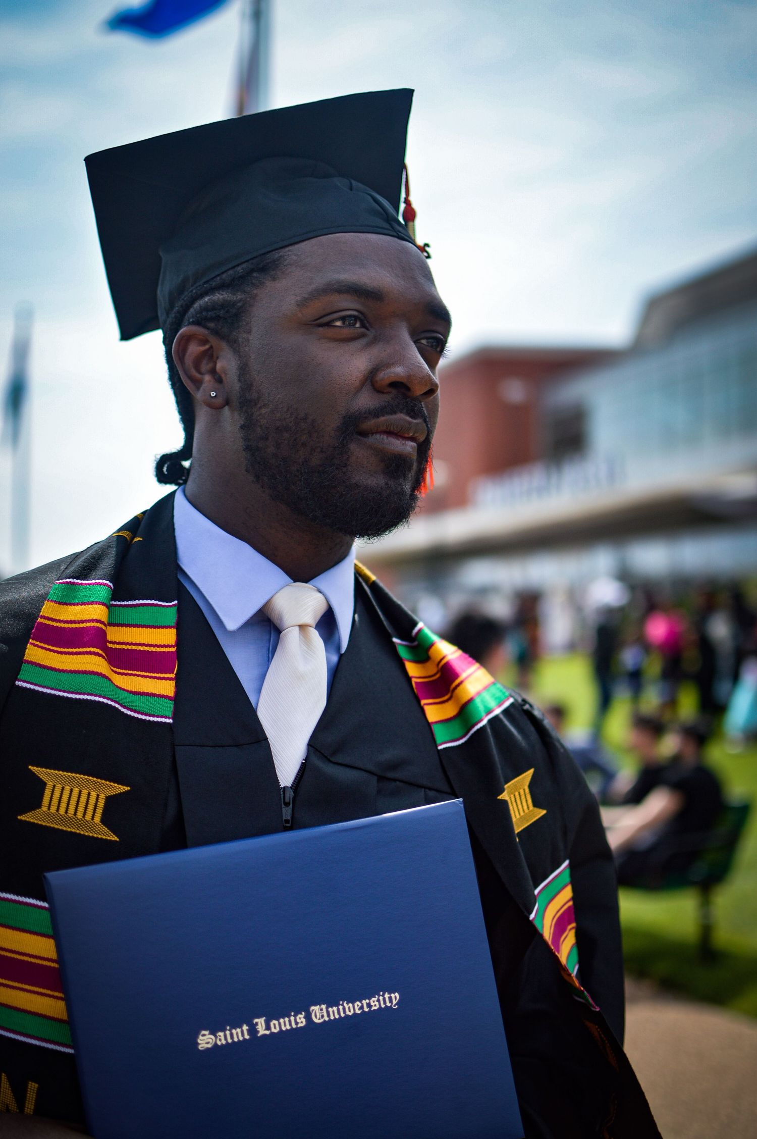 Proud graduate in academic regalia displaying colorful kente stole and diploma during commencement.