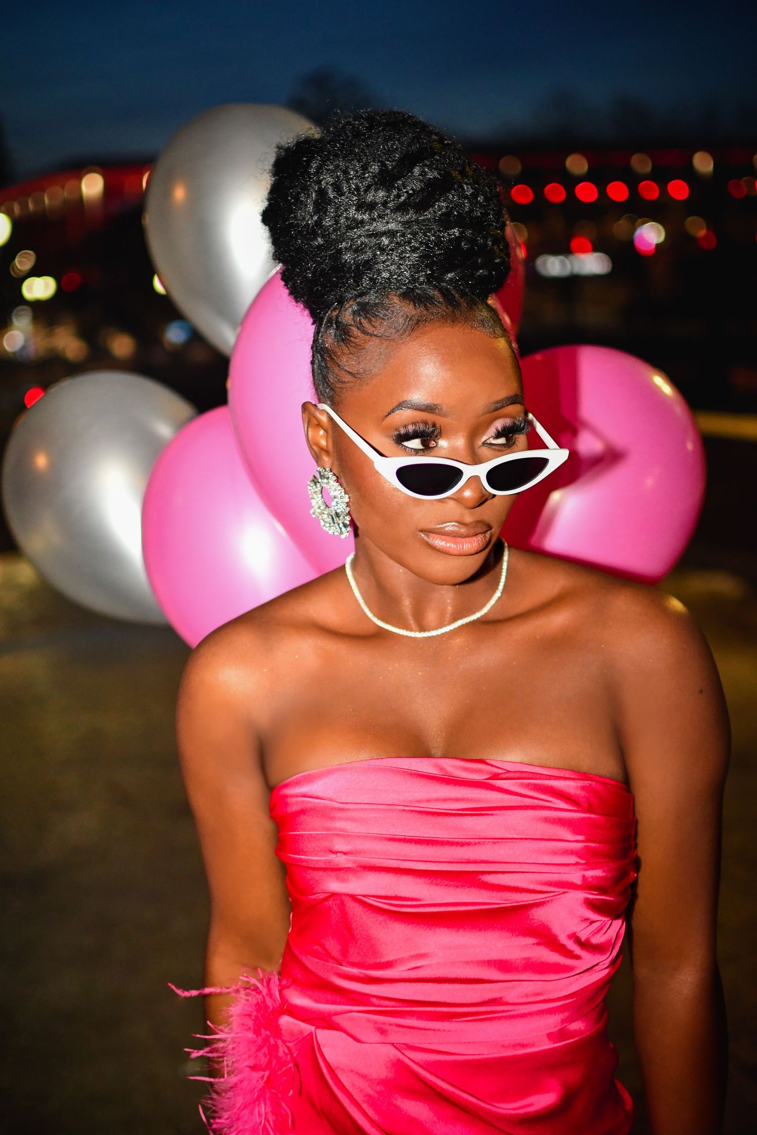 Stylish partygoer in pink strapless dress and white sunglasses poses with silver and pink balloons in nighttime setting.