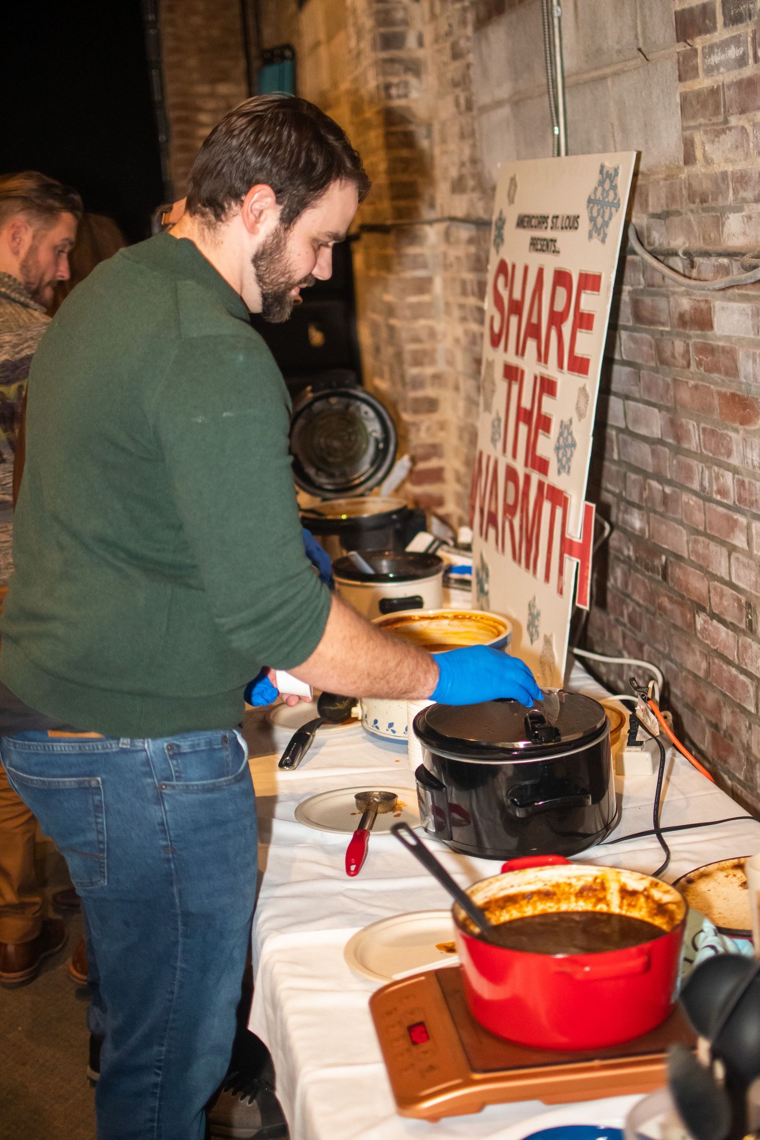 Person in green sweater preparing food at a buffet table with crockpots and serving dishes against brick wall.