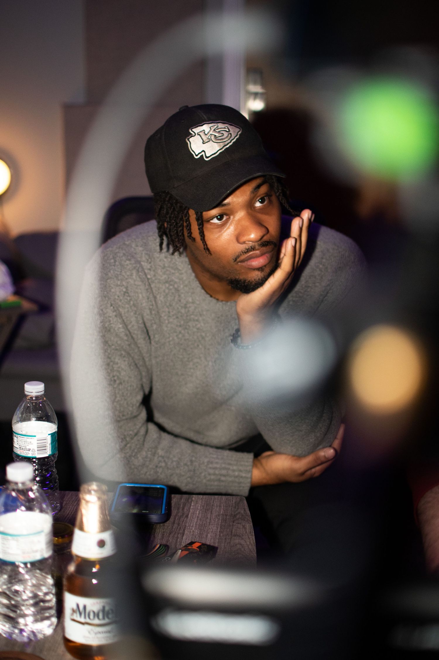 Person in gray hoodie and black hat sits at dimly lit table with water bottles in atmospheric nighttime setting.