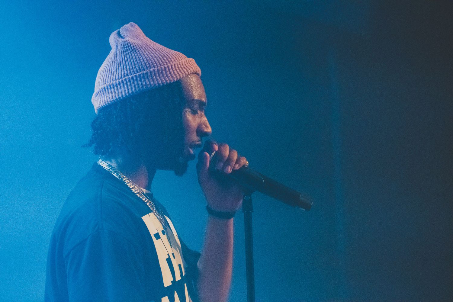 A performer in a white beanie sings into a microphone against a blue-lit stage backdrop.