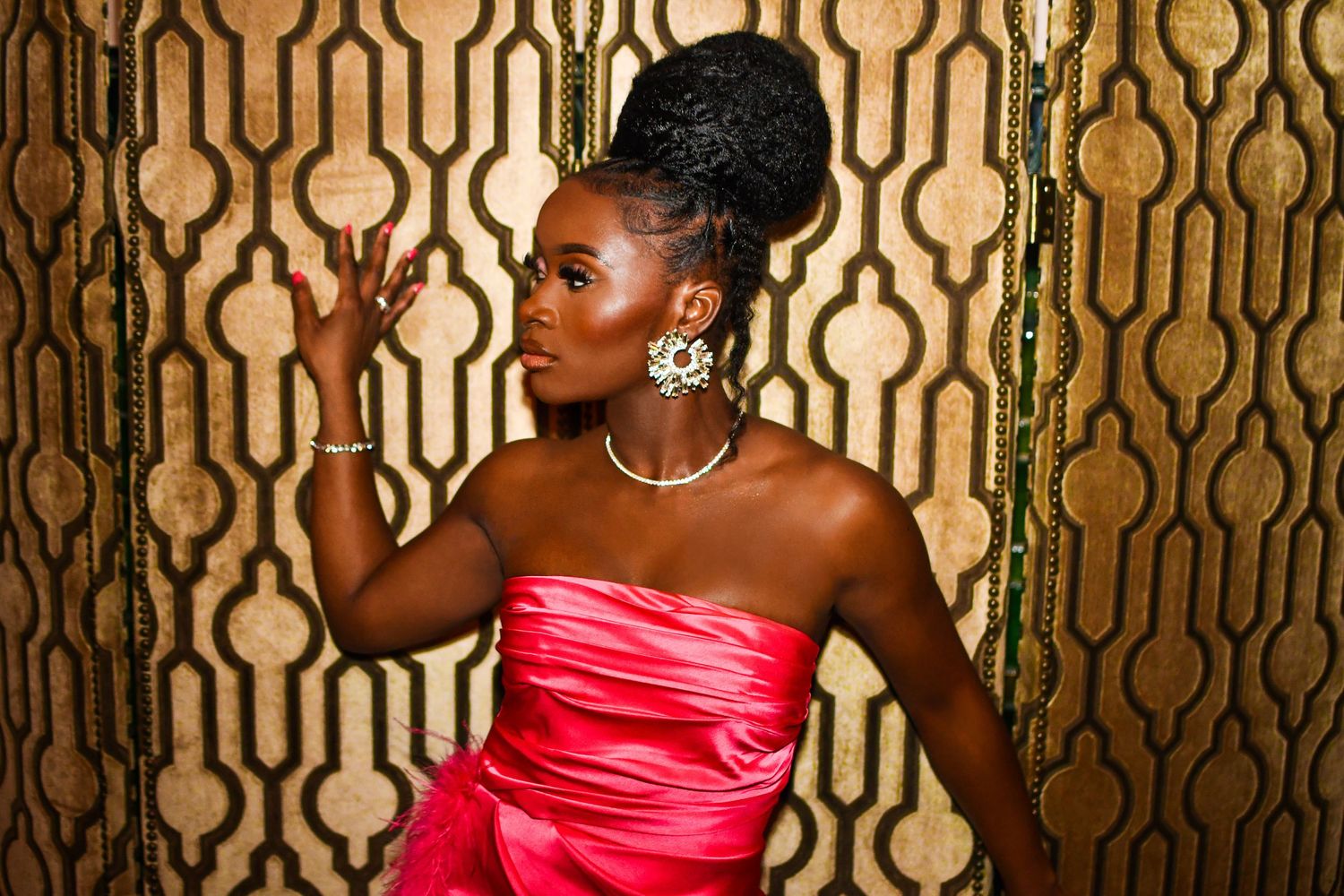 Elegant figure in strapless pink satin gown poses against gold patterned wallpaper wearing sparkling statement earrings.