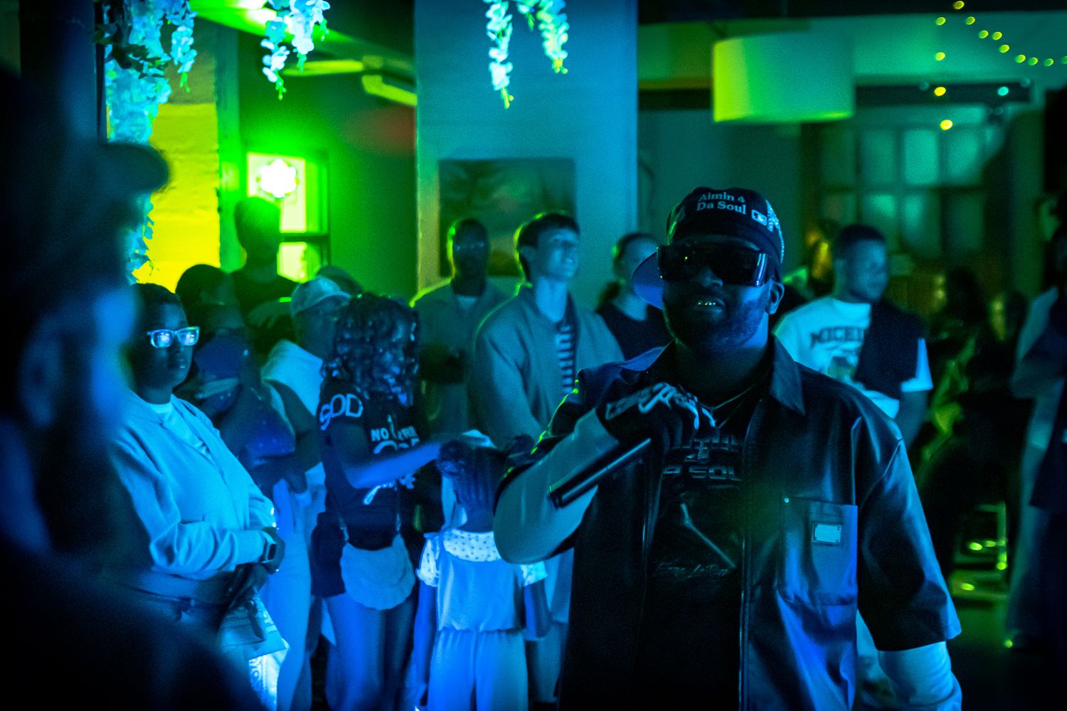 A performer holds a microphone at a vibrant green-lit nightclub event with a crowd in the background.