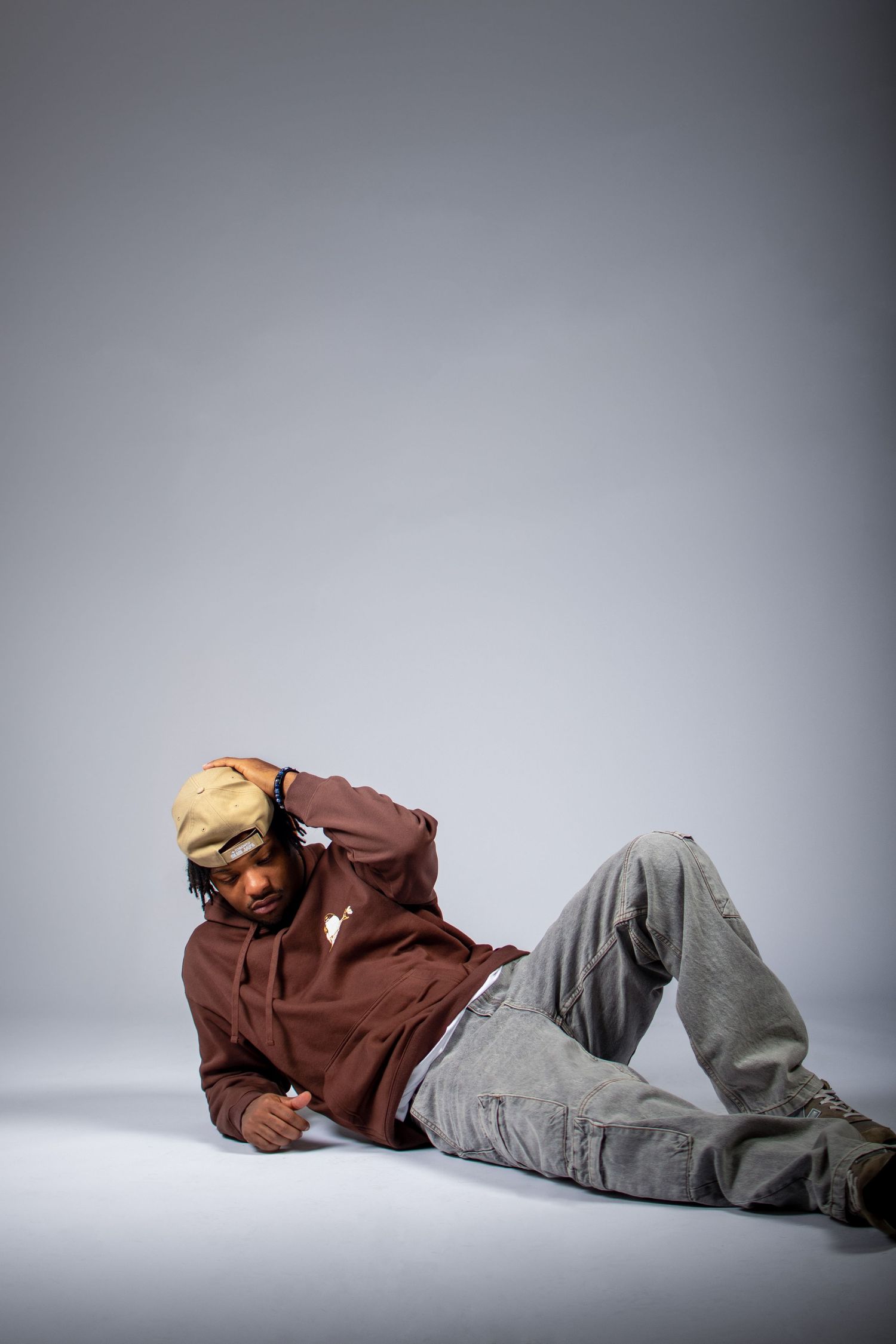 A person in a brown hoodie and gray pants lies on their side against a light gray background in a dramatic pose.