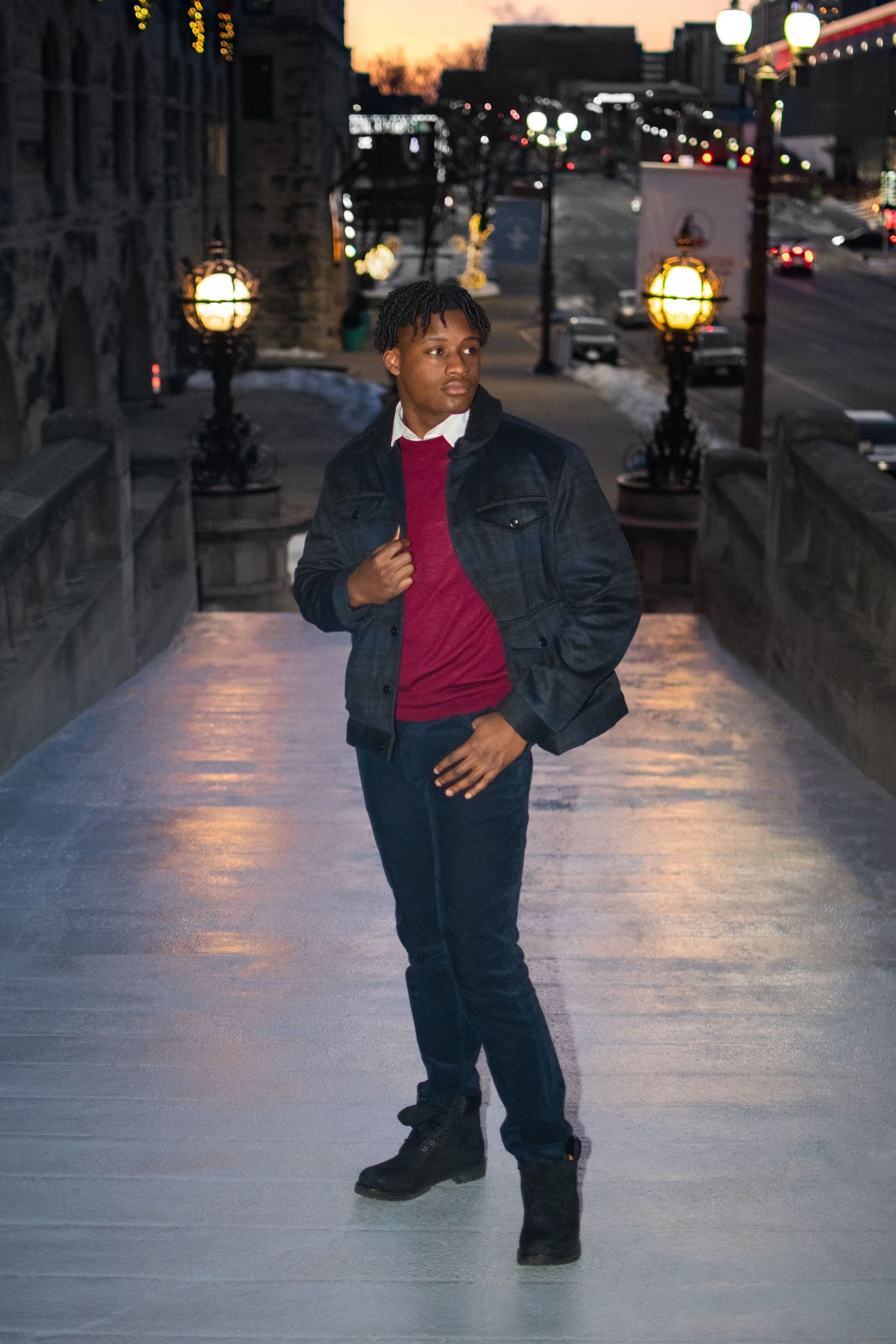 A person poses on an illuminated walkway at dusk wearing a denim jacket, burgundy sweater and dark jeans.