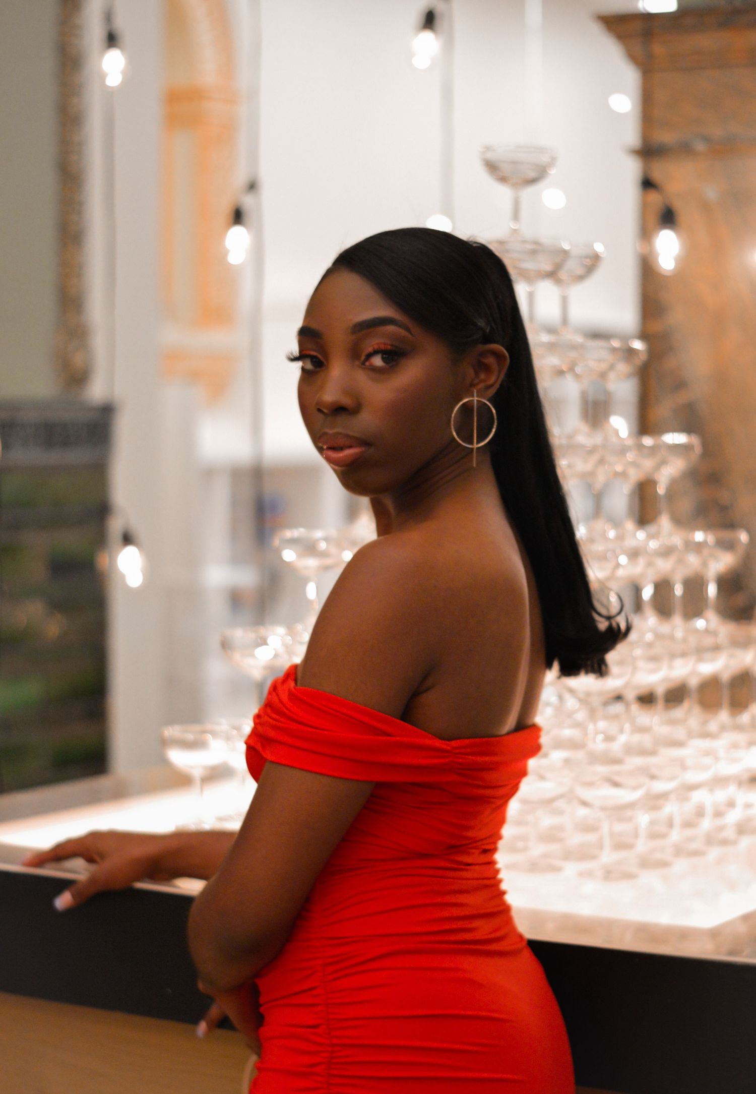 Woman in a vibrant red off-shoulder dress poses elegantly against a backdrop of glowing pendant lights.