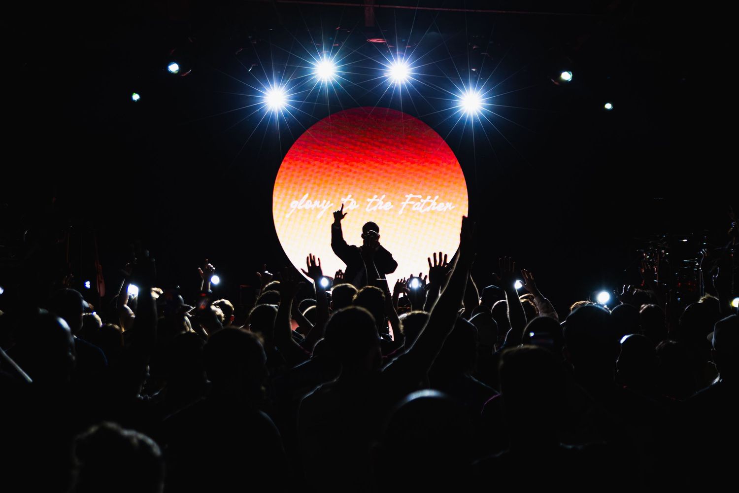 Silhouetted performer on stage with glowing orange circle backdrop and crowd raising hands at a live concert.