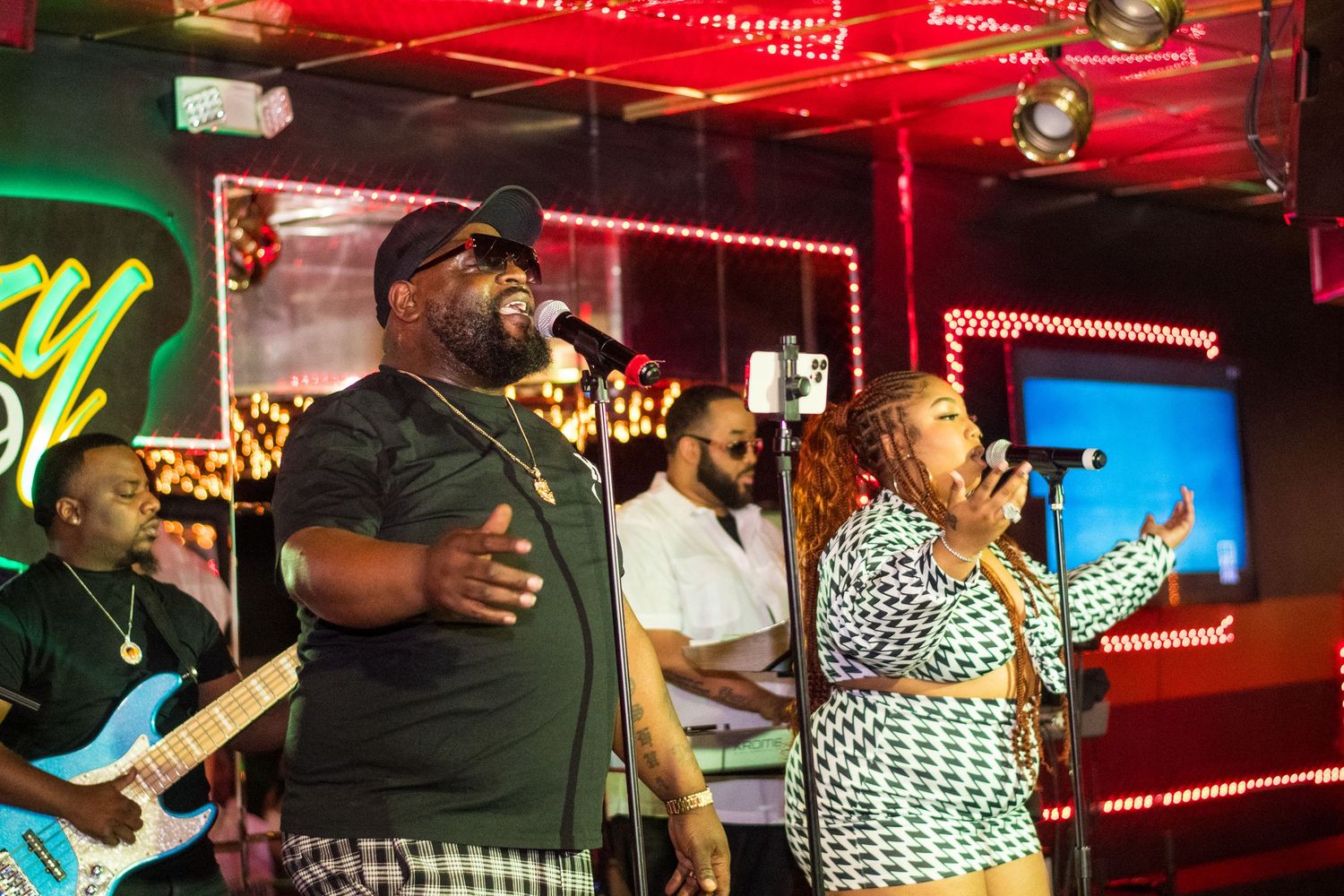 Musicians perform energetically on stage at a club with red neon lighting and band equipment visible in the background.