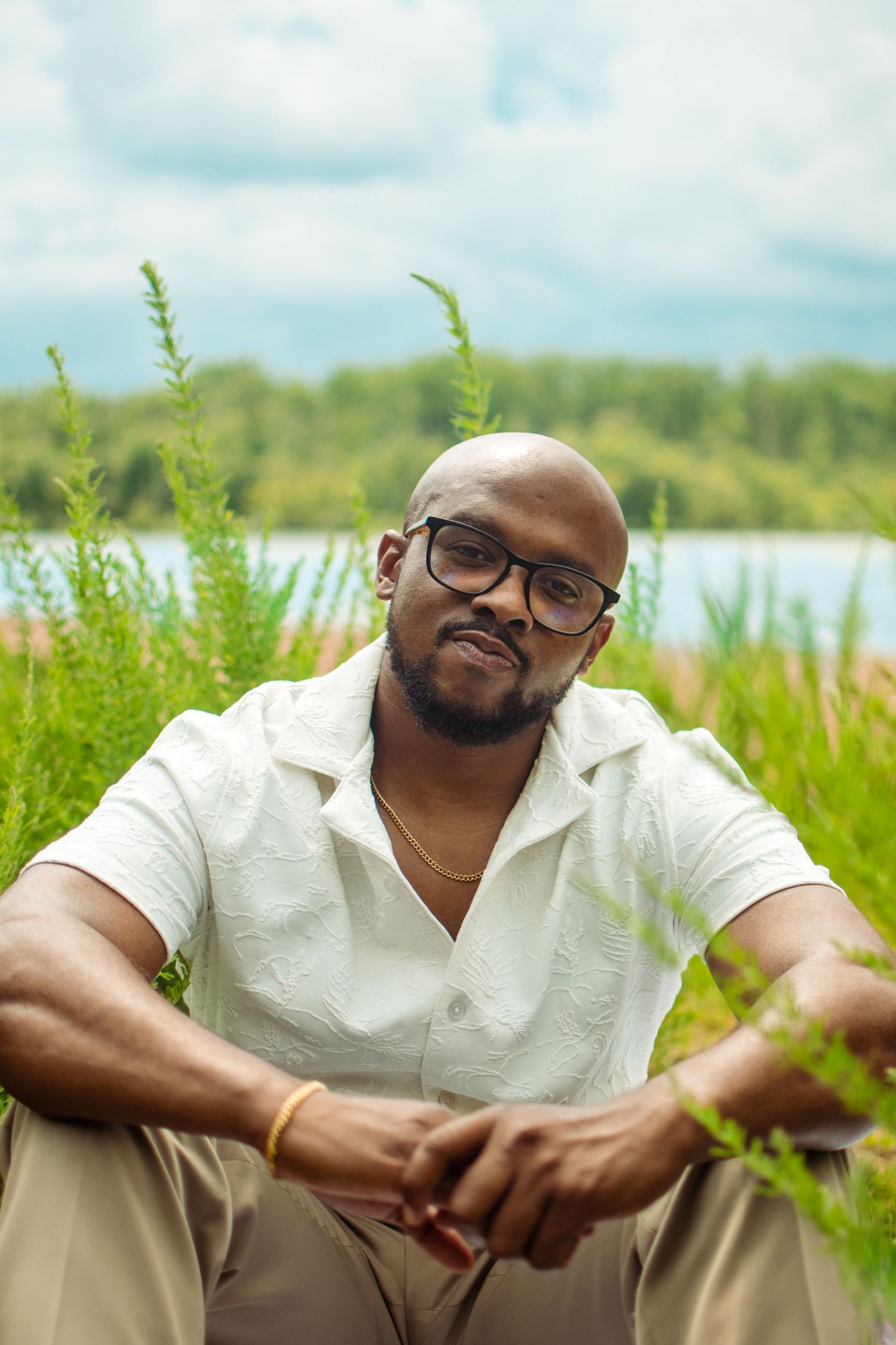 A person wearing glasses and a white polo shirt sits outdoors near a lake with tall grass and trees in the background.