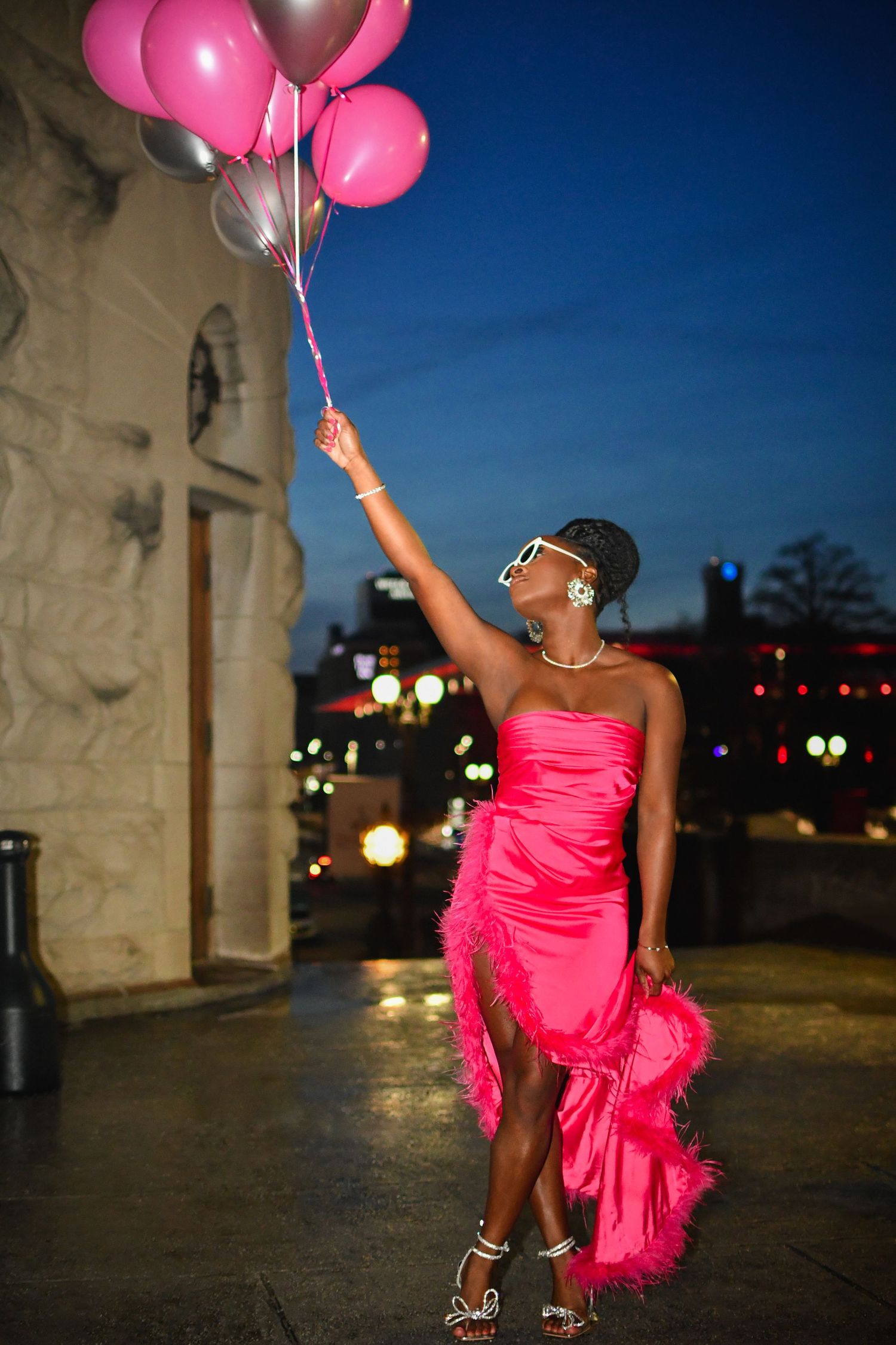Person in a bright pink dress dances with balloons against a dark blue evening sky with city lights.