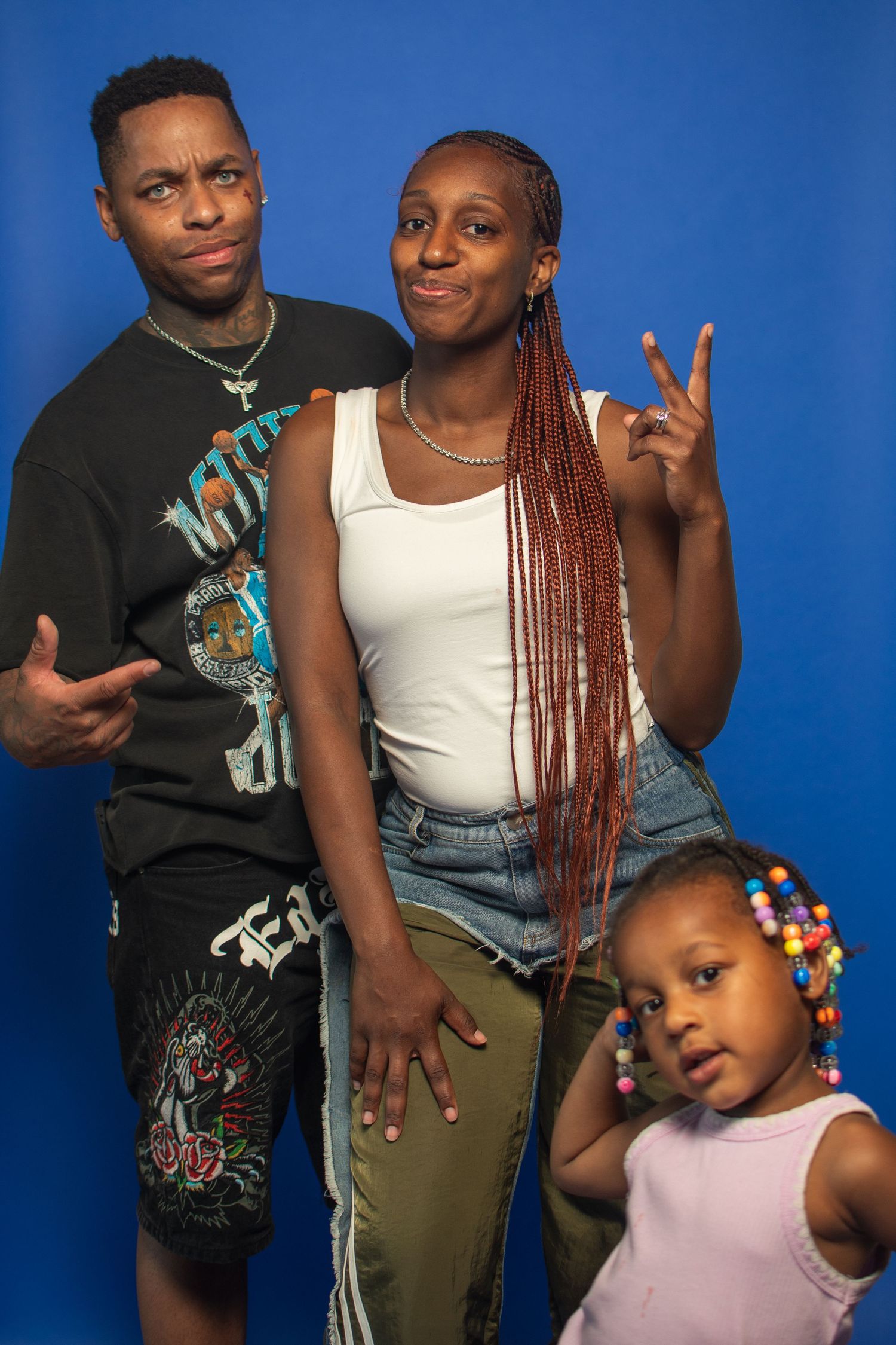 Three people pose together against a blue backdrop, with one wearing a graphic tee, another in a white tank top, and a child in pink.