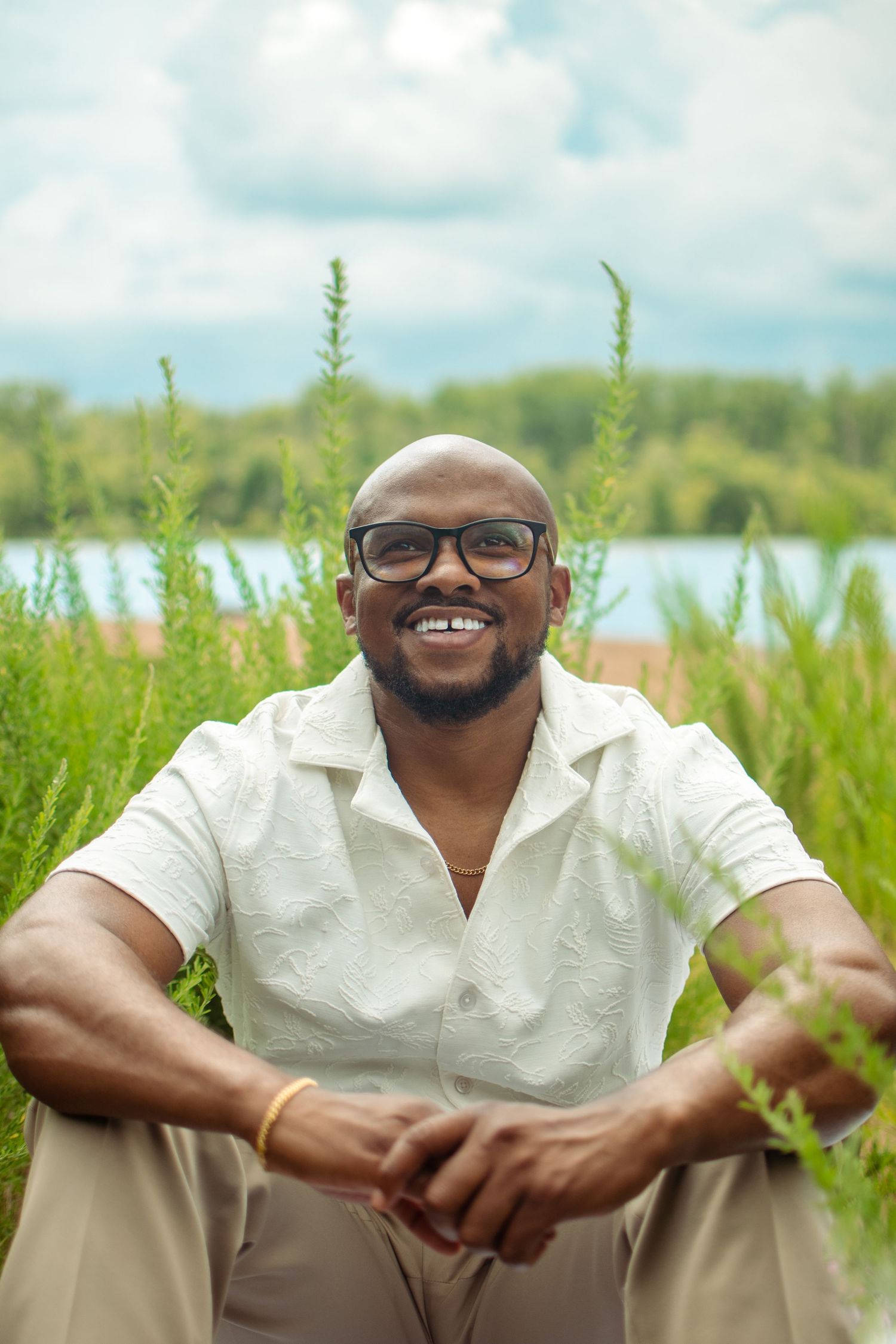 A person in a white shirt sits outdoors in a field of tall grass with a bright smile and glasses.
