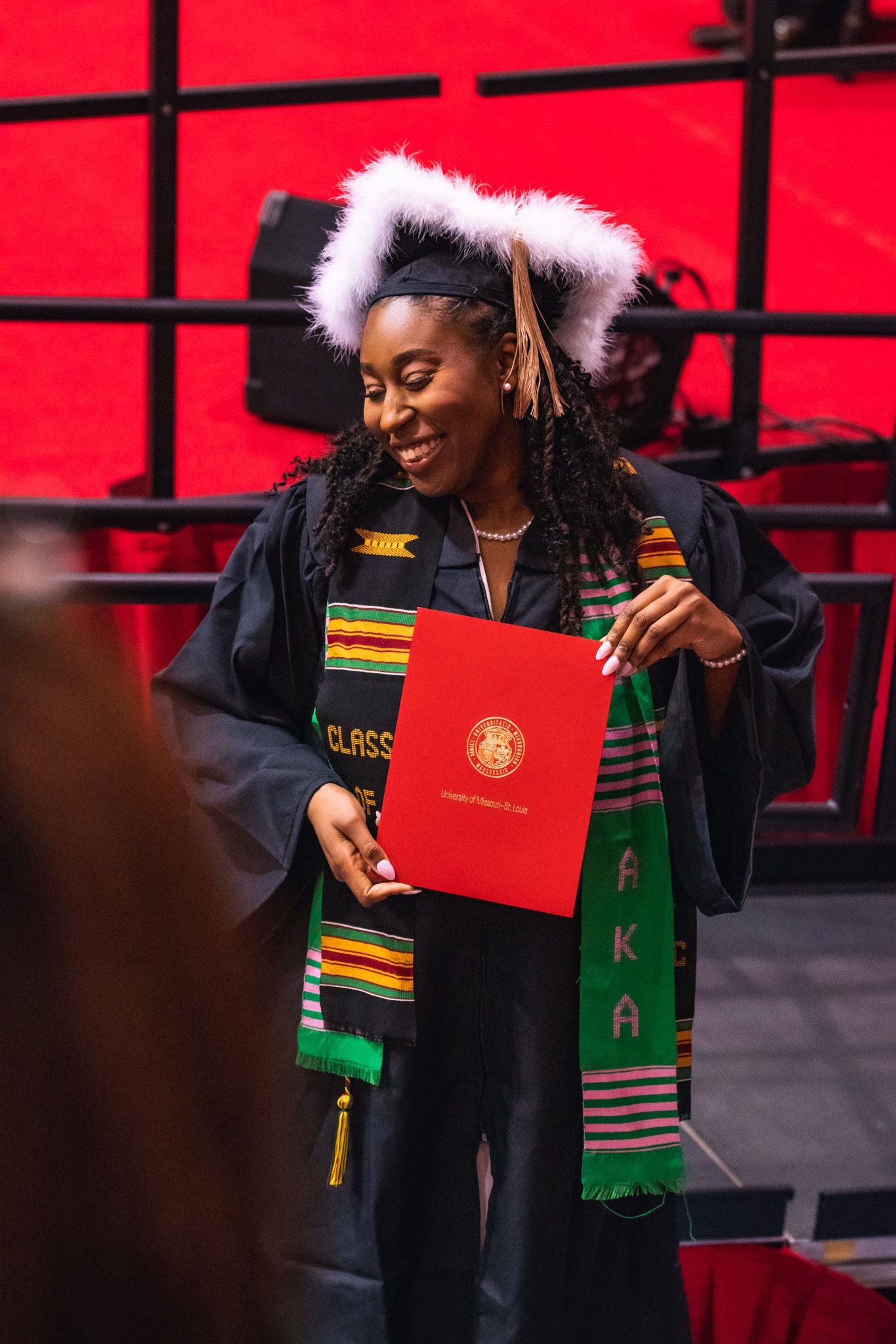 A graduate in black cap and gown with kente stole smiles while holding a red diploma during commencement ceremony.