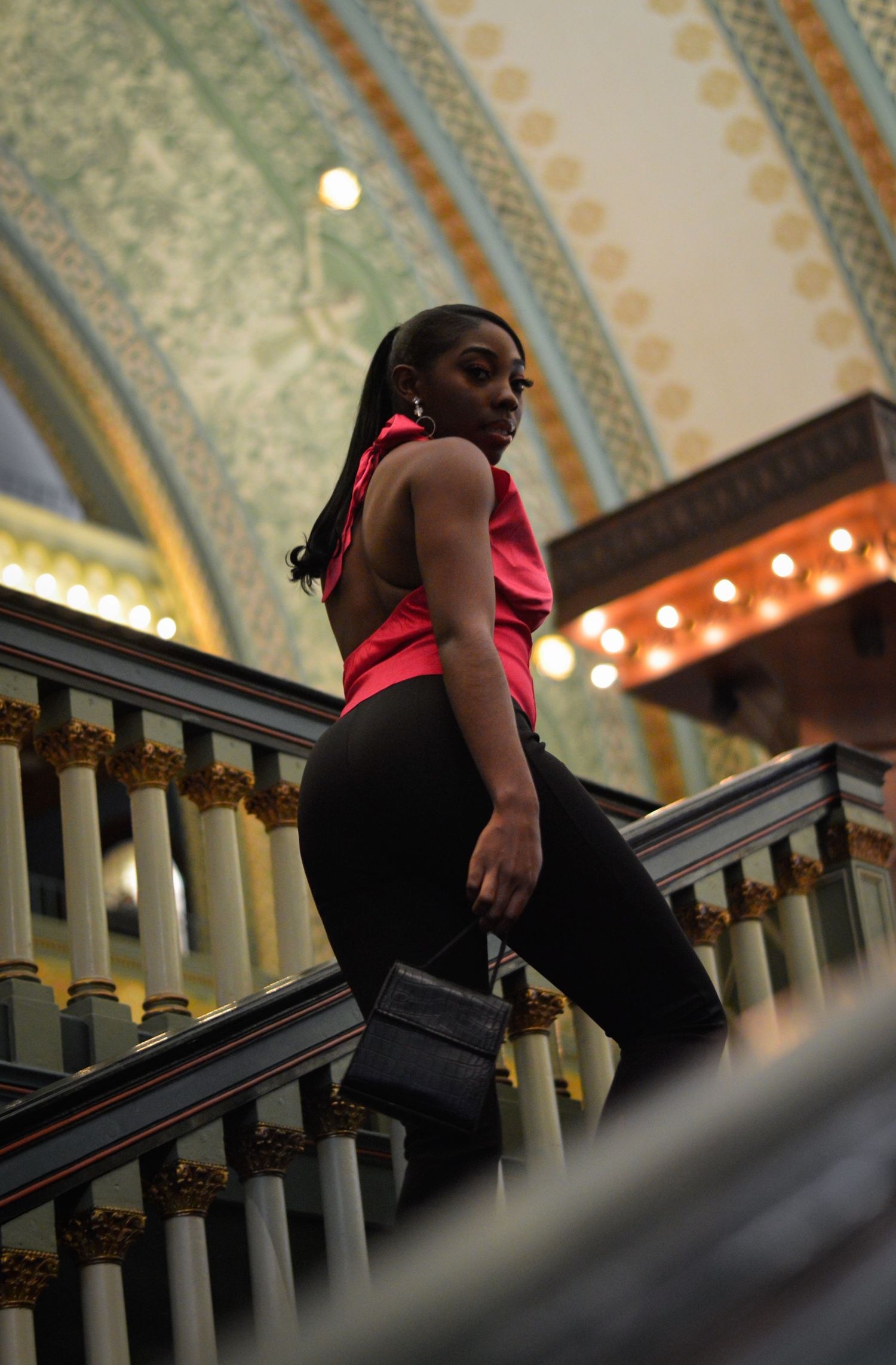 A black woman in black and pink attire descends an ornate staircase with decorative railings under a patterned ceiling.