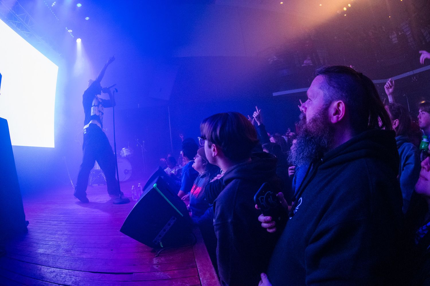 Concert audience members watch a performer on stage in a dark venue with dramatic blue lighting and fog effects.