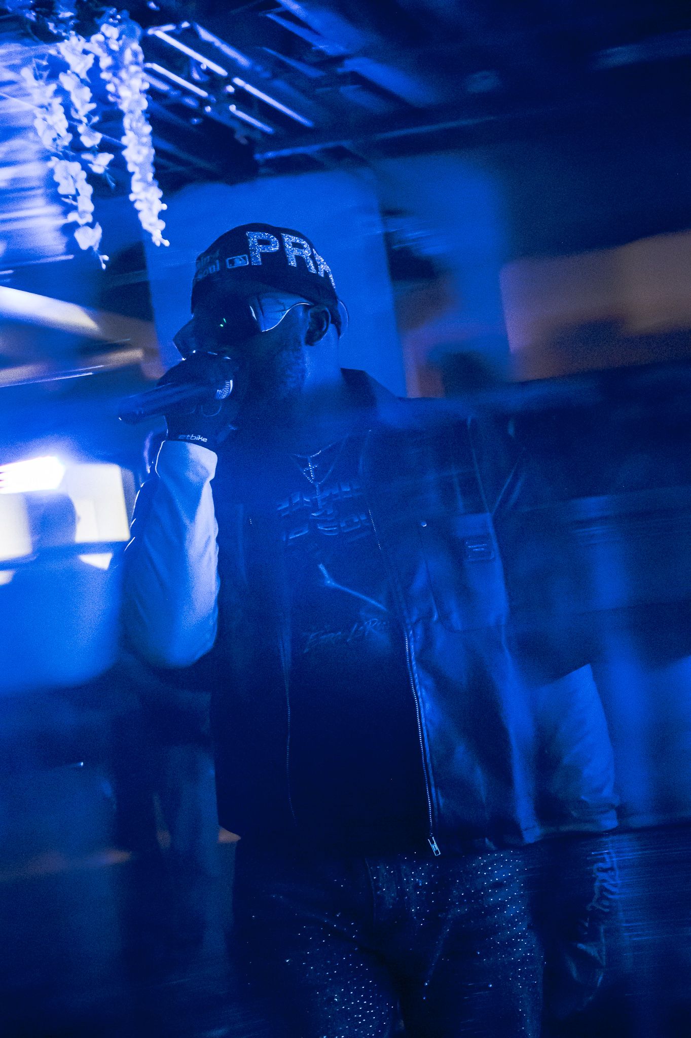 Performer rapping into microphone under blue stage lights wearing a PRA cap at a nightclub.