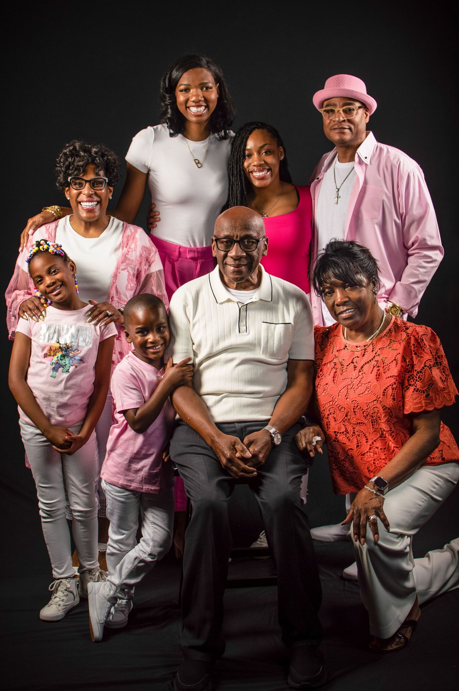 Multi-generational family wearing pink and white outfits poses together against dark background in studio portraits.