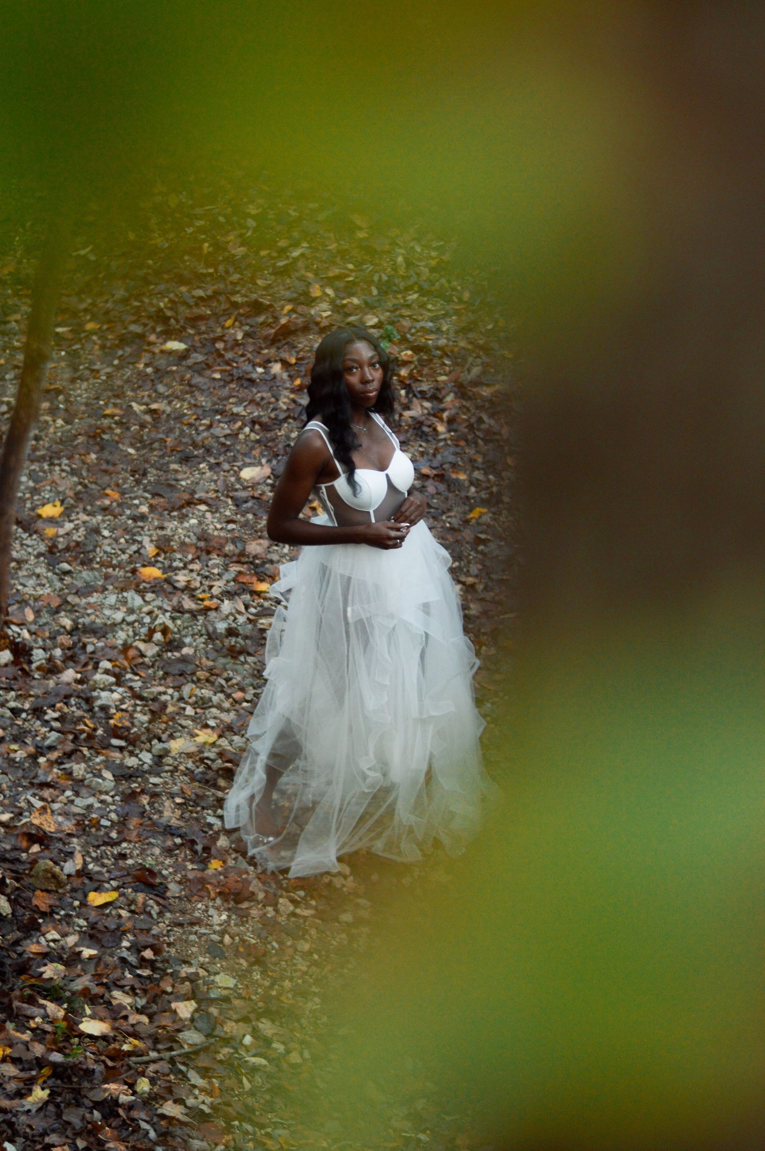 Two children in white dresses sit together on fallen autumn leaves in a wooded outdoor setting.