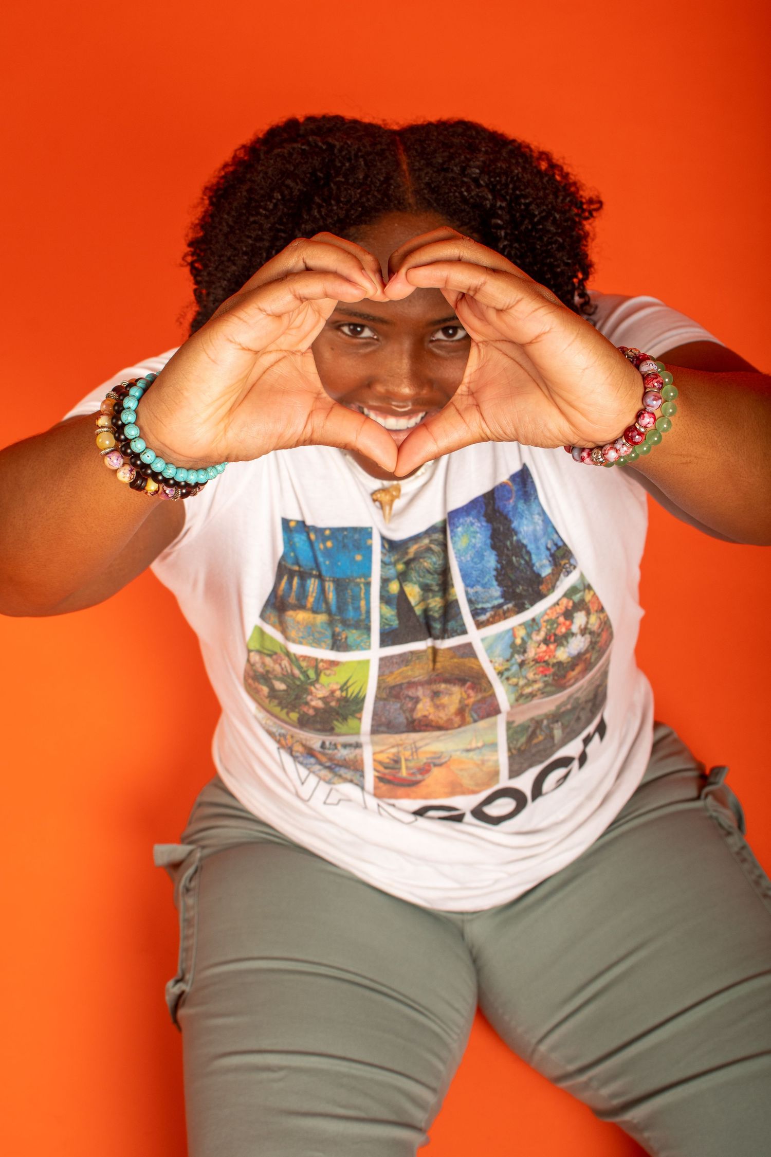 Person in gray pants and printed t-shirt poses playfully against orange background making heart shapes with hands.