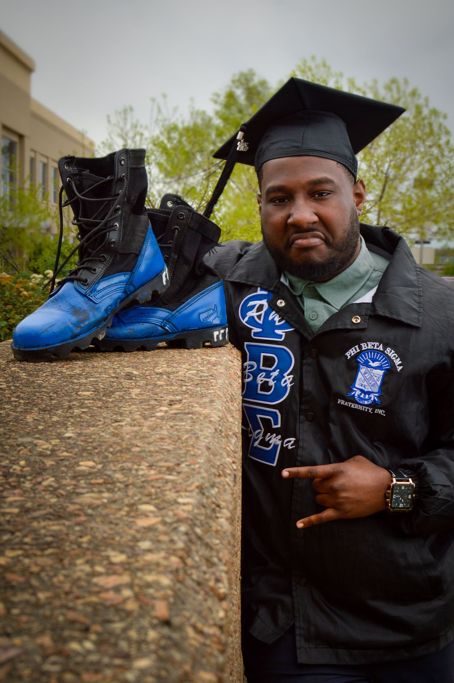 Graduate in black gown displays diploma while sitting on stone wall outdoors.
