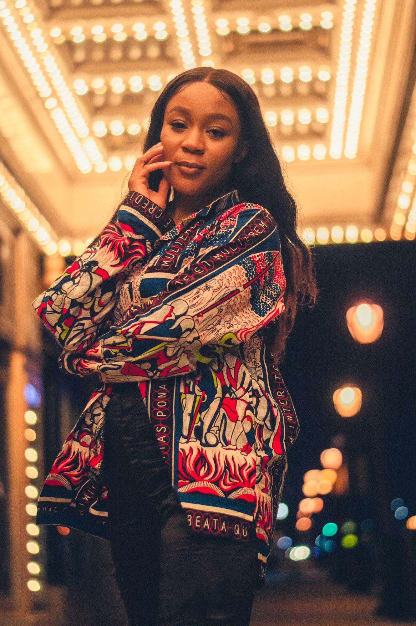 A fashionable portrait against illuminated lights wearing a colorful patterned bomber jacket and black pants.