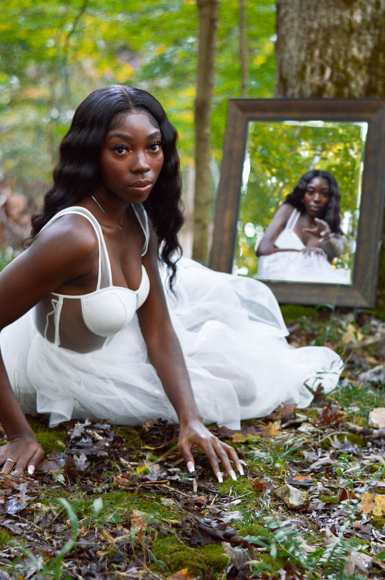 A person in a white dress lies gracefully on autumn leaves in a wooded outdoor setting.