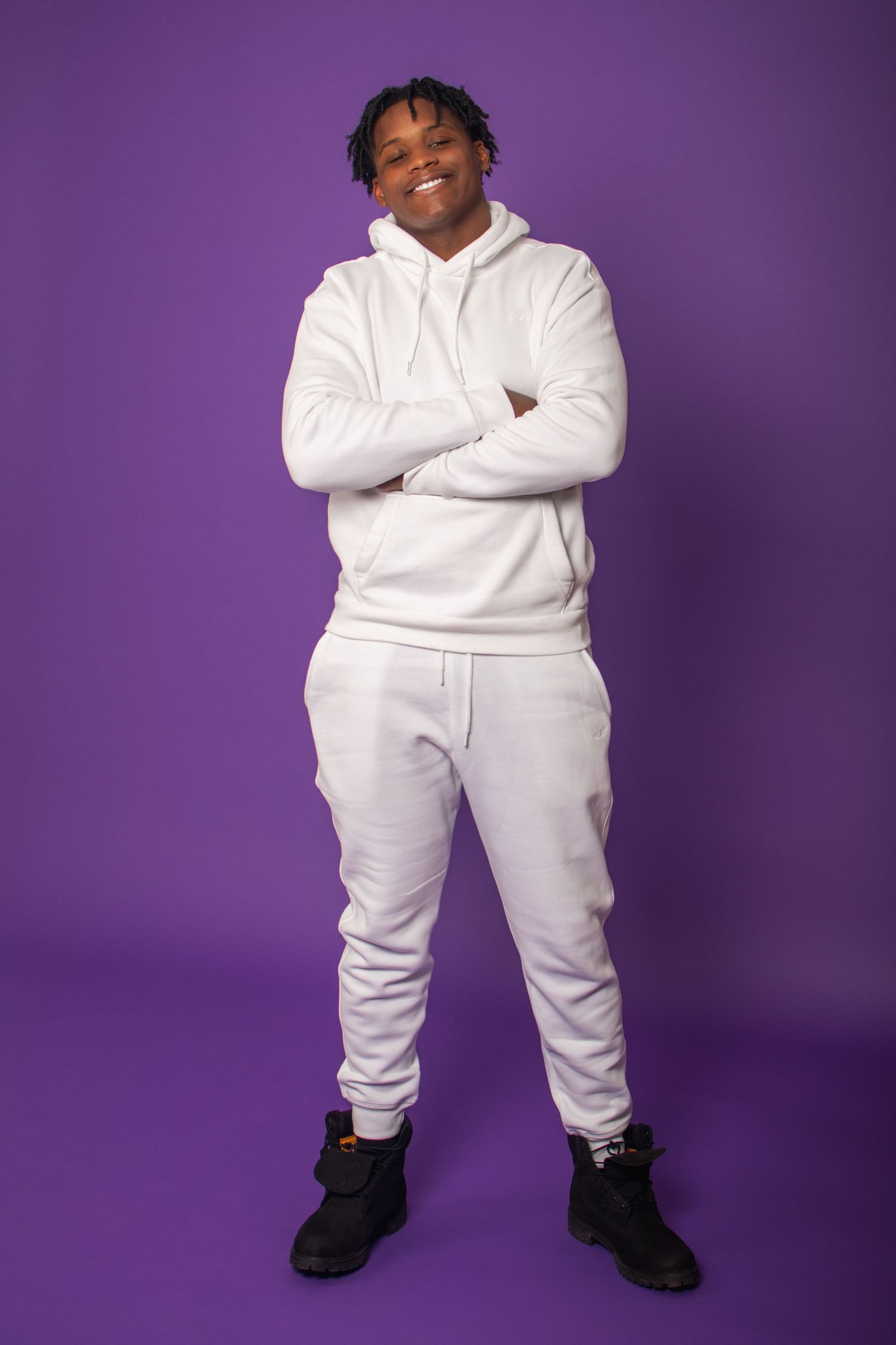 Person wearing all white sweatsuit and black shoes poses against purple backdrop in multiple poses.