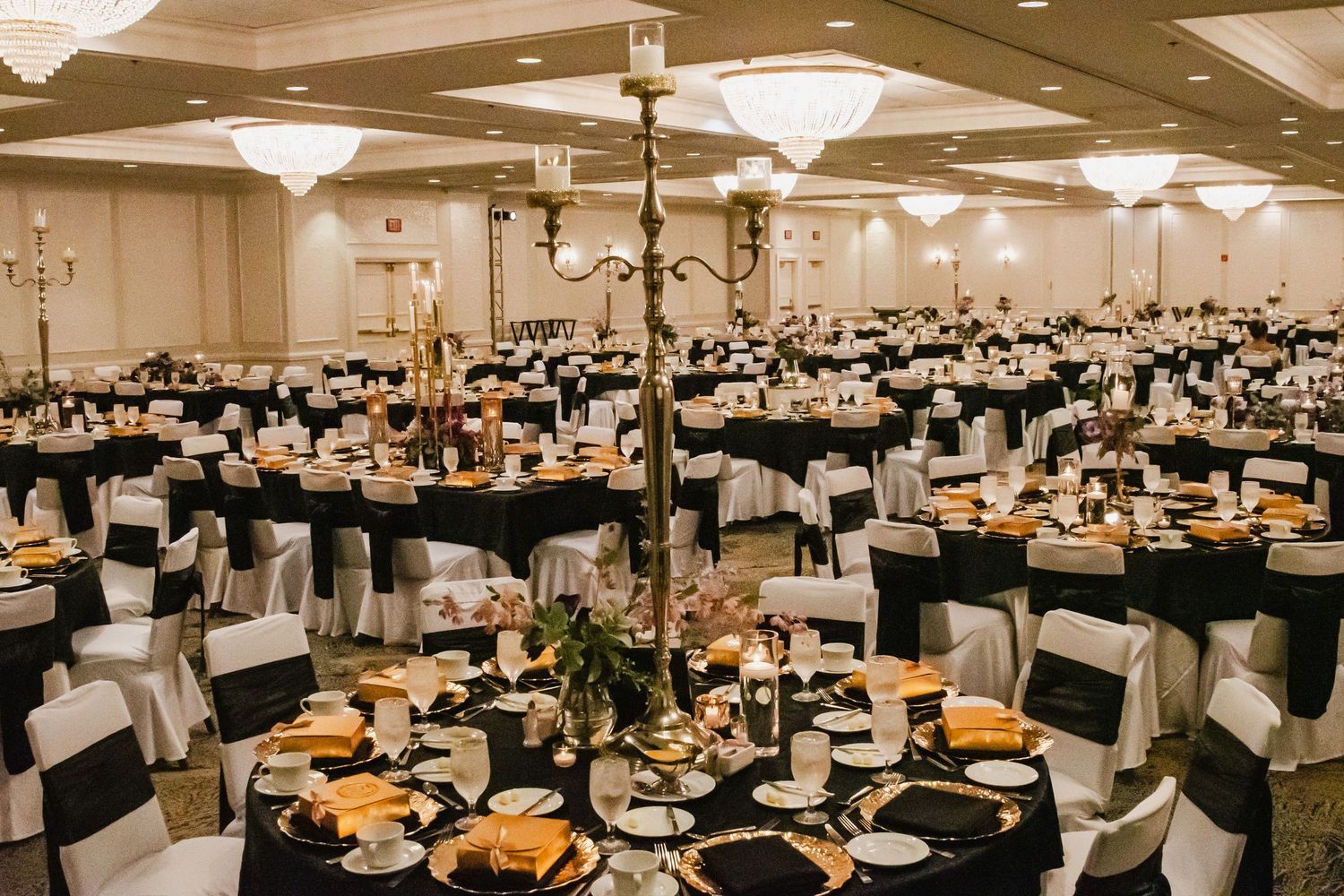 Elegant ballroom with black and white chair covers, gold accents, candelabras, and round tables set for a formal event.