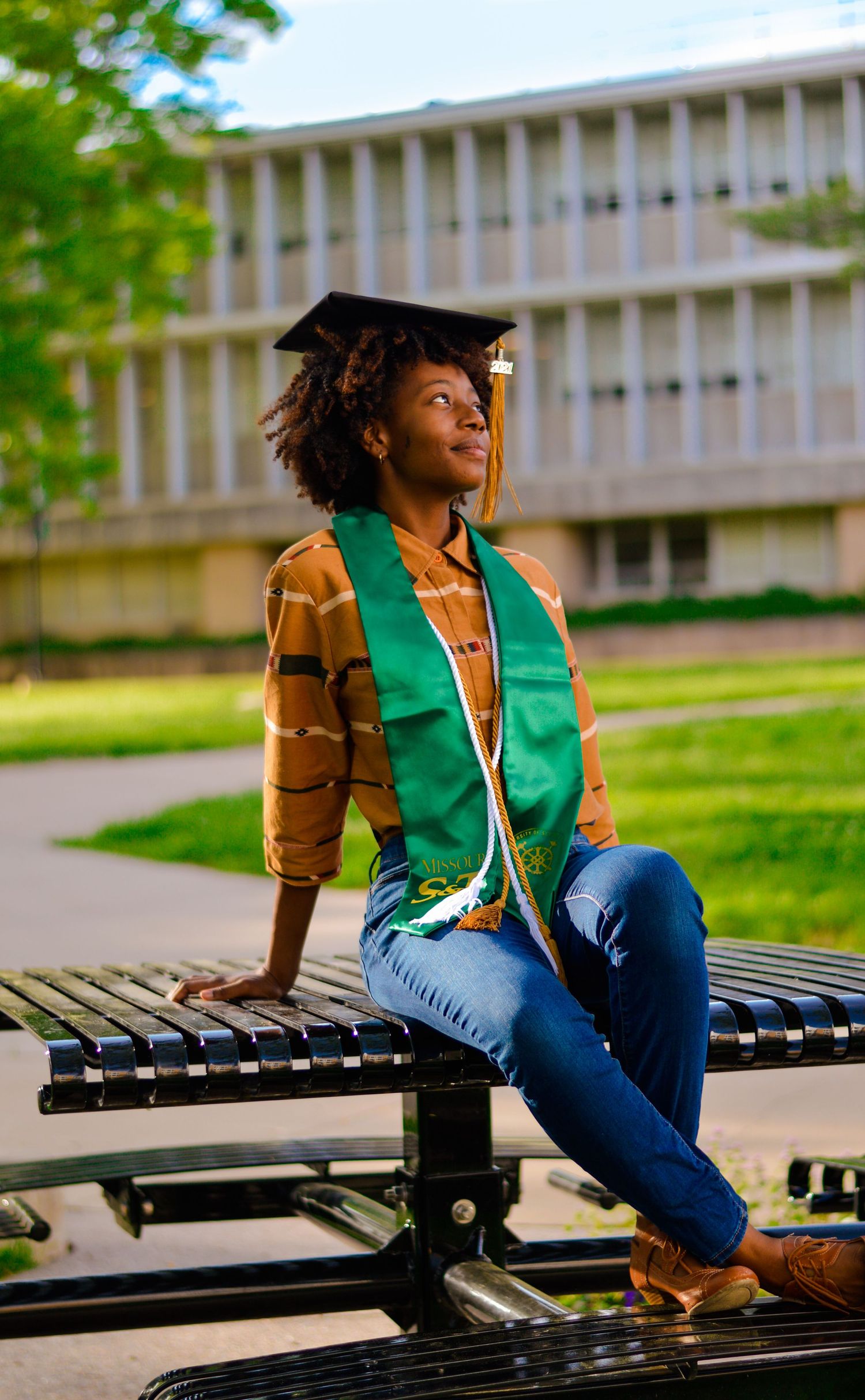 A black woman in a green vest and brown jacket sits casually on wooden benches outside a modern building.