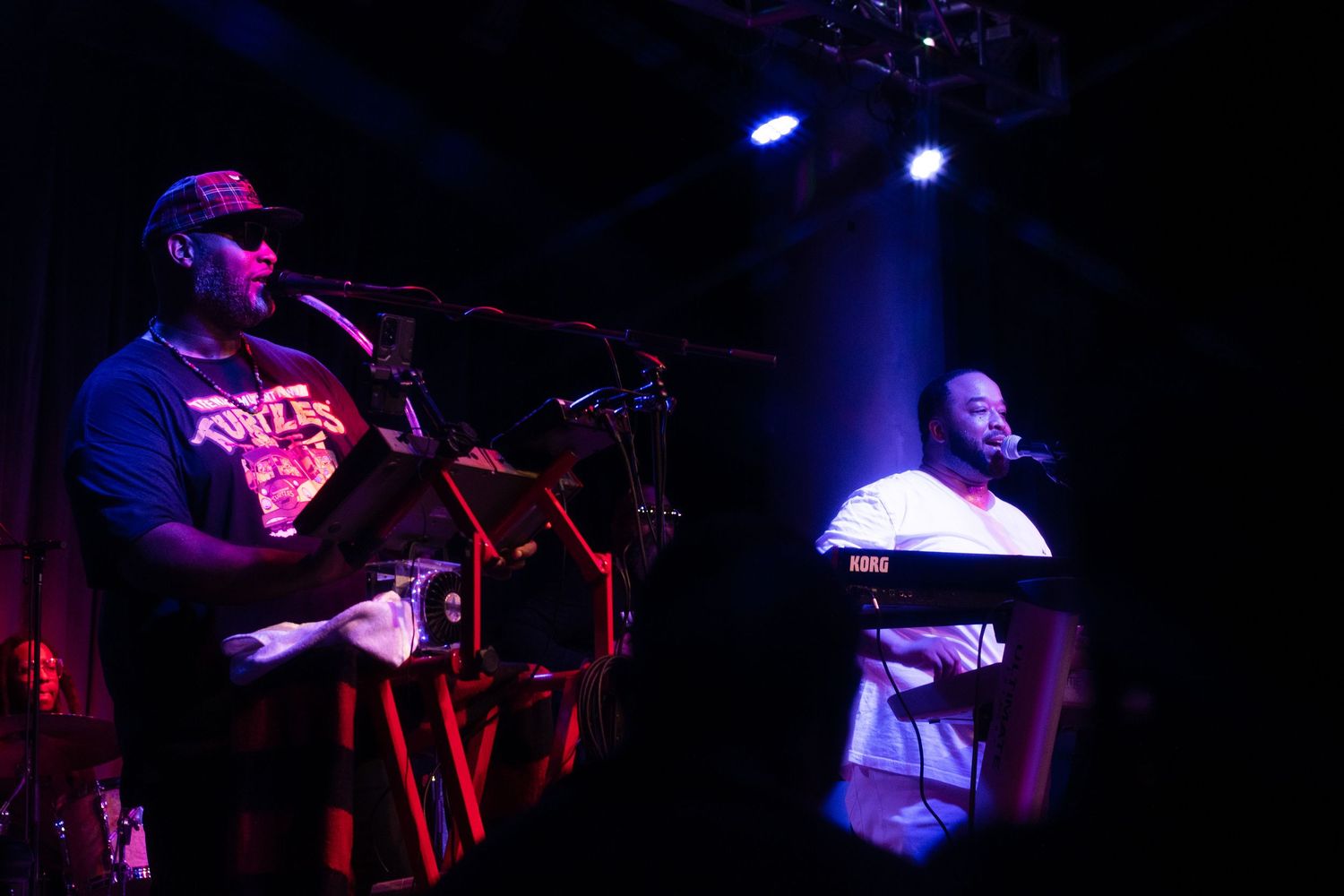 Musicians perform on stage under blue lighting at a concert venue with one person singing and another at a keyboard.