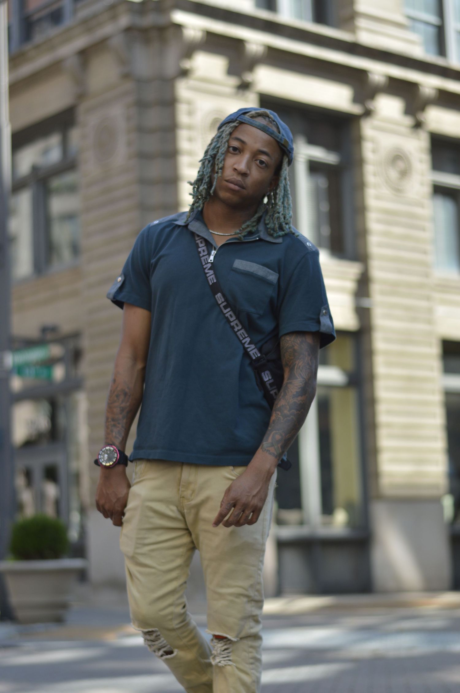 Person in dark teal shirt and khaki pants poses casually on urban street with architectural backdrop.
