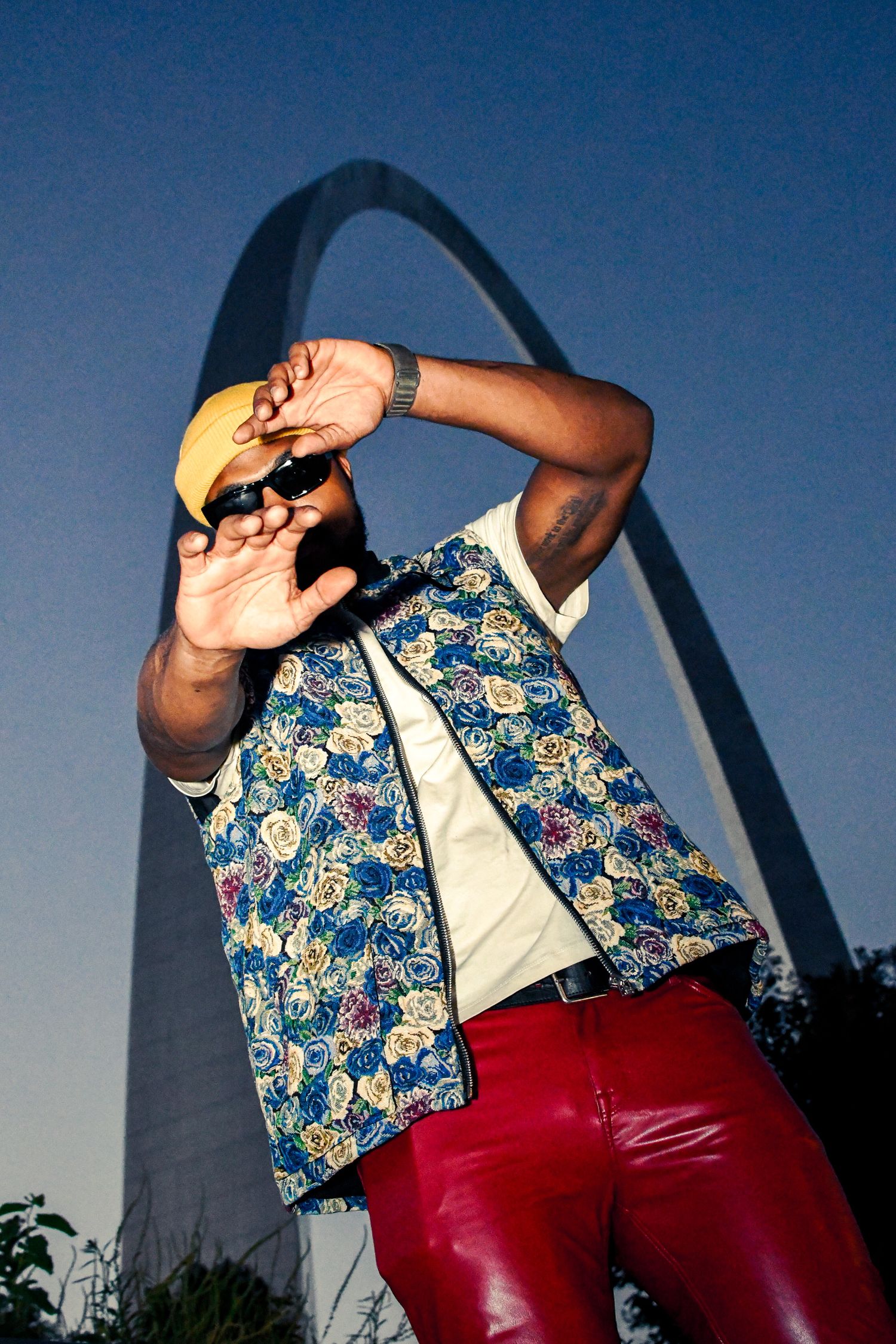 A person wearing red pants and a floral vest poses in front of the Gateway Arch in St. Louis at dusk.