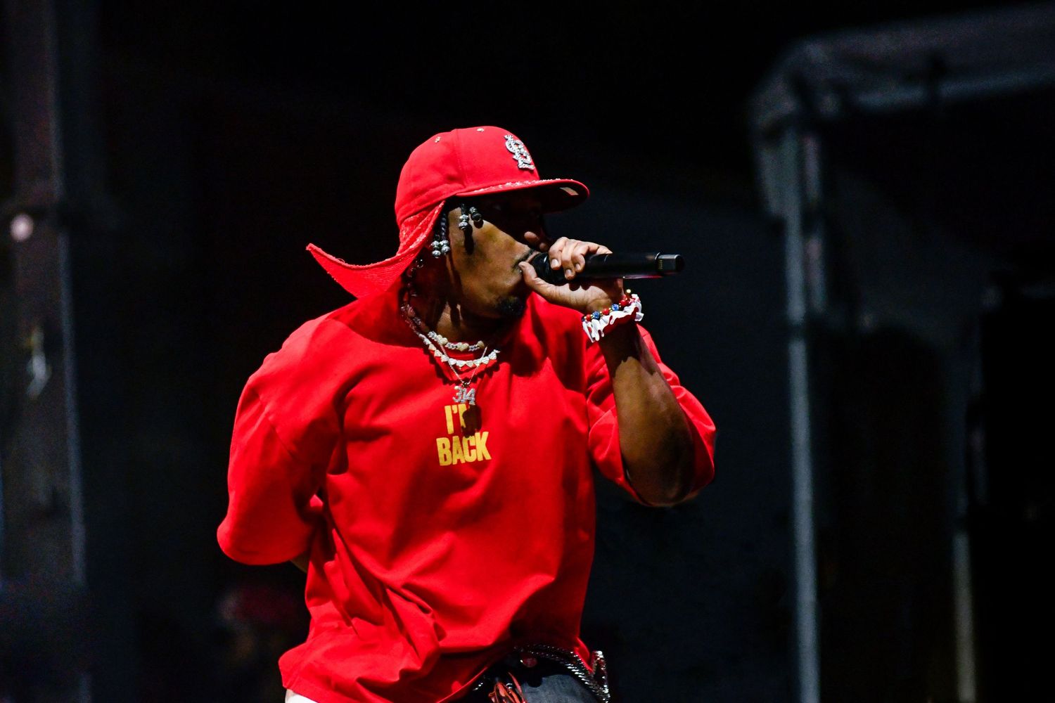 Performer in bright red outfit and baseball cap holds microphone while performing on stage during concert.