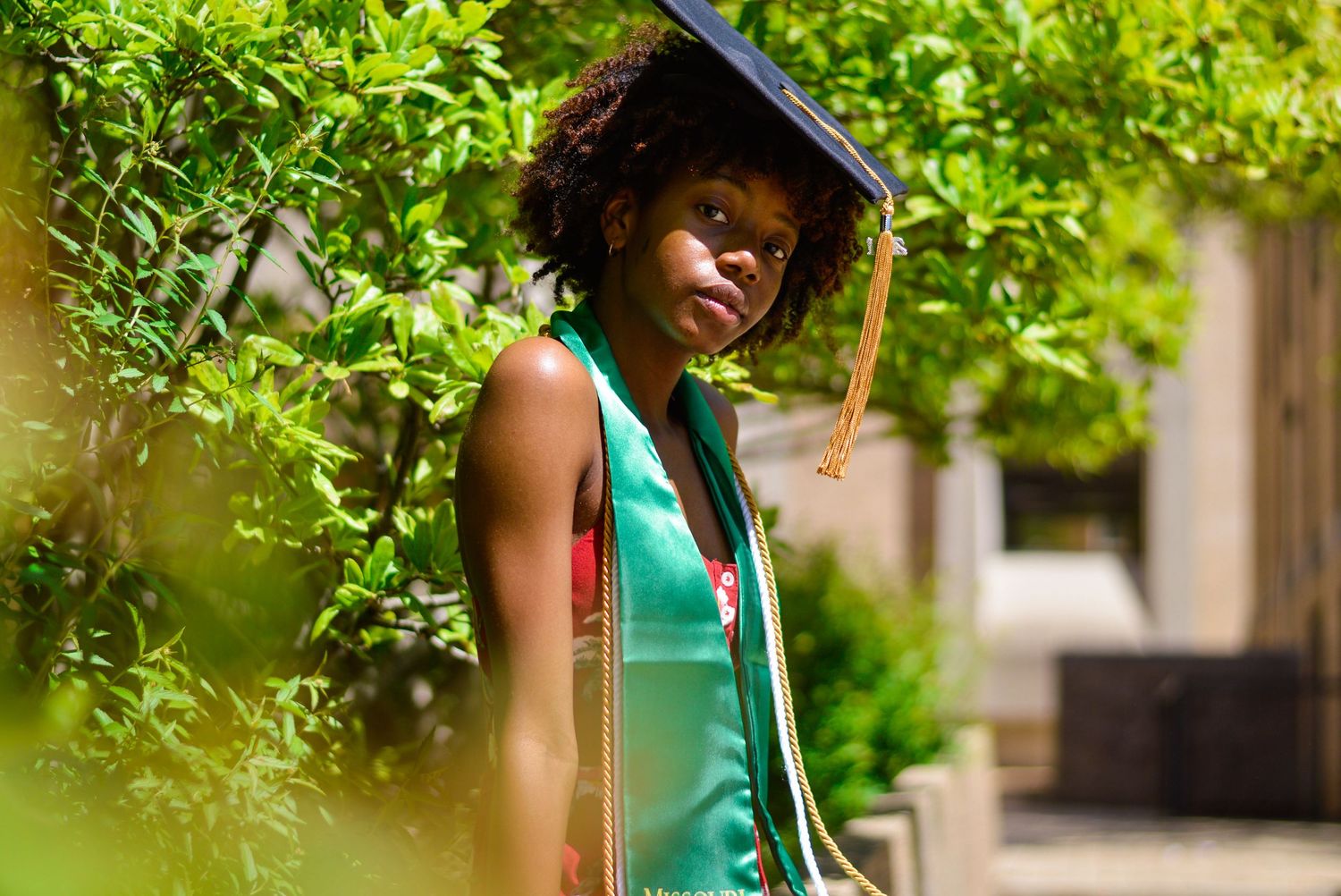 Graduate in emerald green dress poses outdoors with greenery in background.