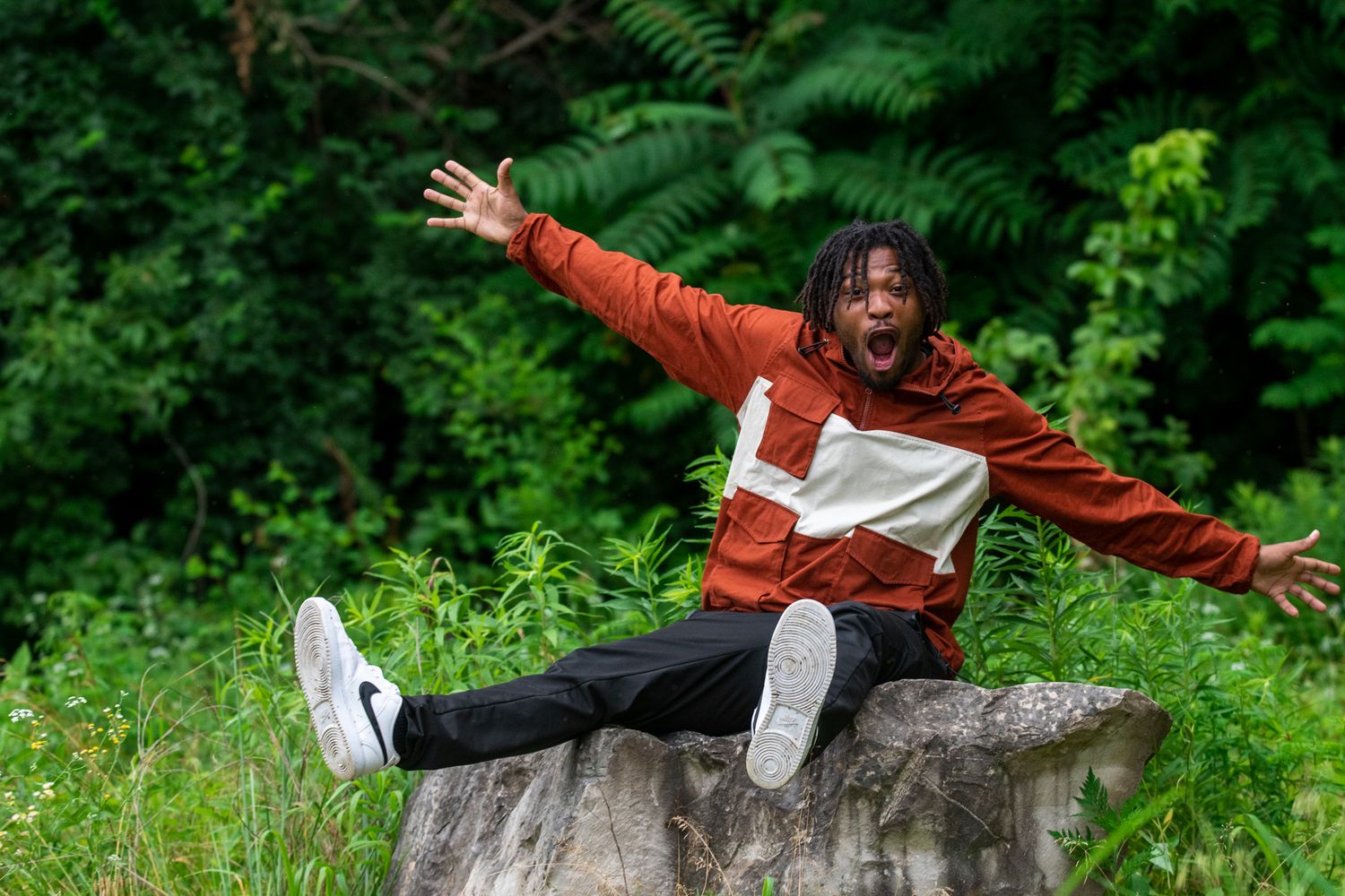 Person in orange jacket and white pants performs dynamic poses on stone wall surrounded by lush green foliage.